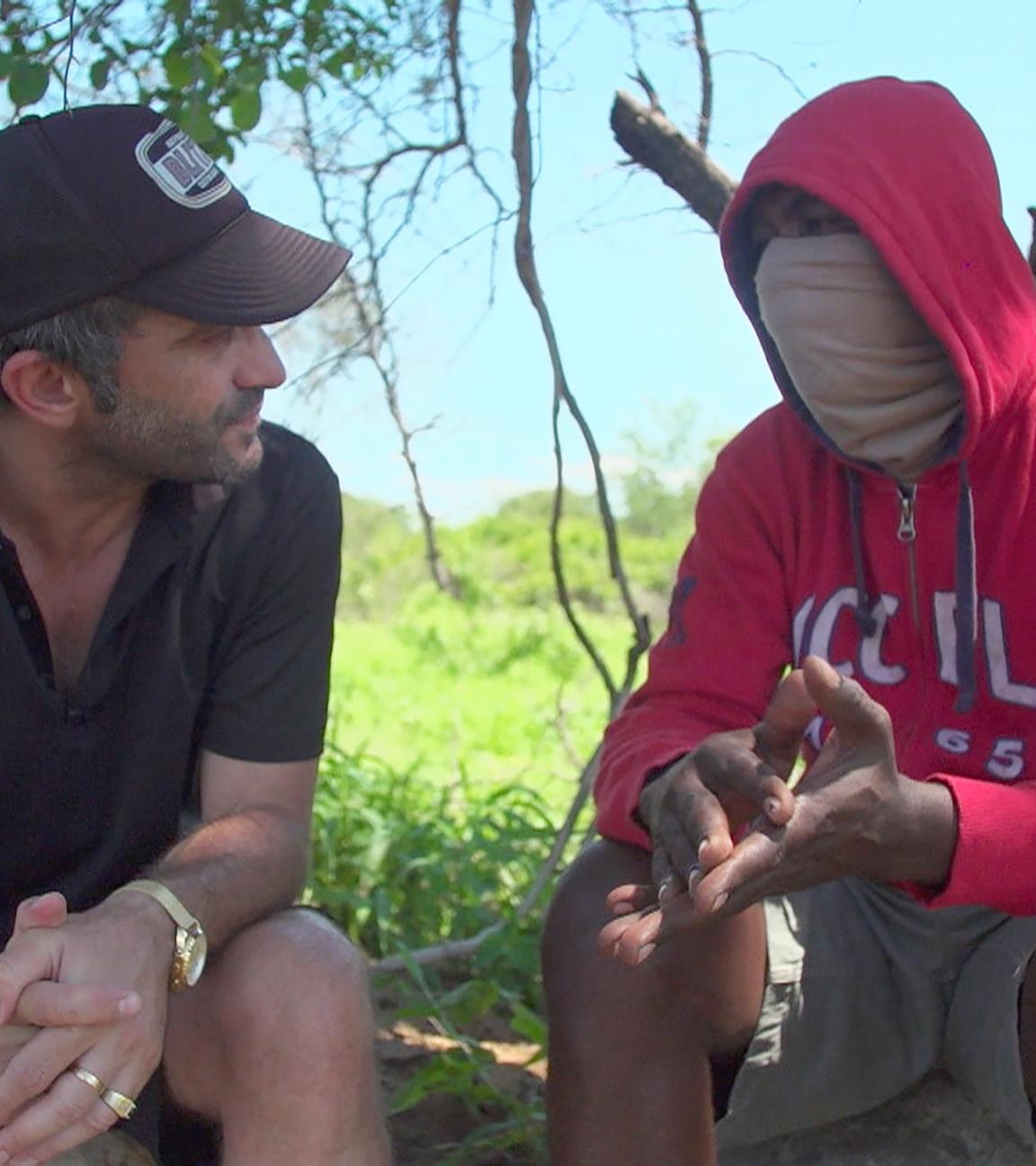 Sebastian Perez Pezzani sitzt mit einem ehemaligen Wilderer im Schatten von einem Baum. Er sitzt links und trägt ein schwarzes, kurzärmliges Hemd und eine schwarze Baseballkappe. Er hat kurze, graumelierte Haare und einen Dreitagebart. Der Mann rechts trägt einen roten Kapuzenpulli mit weißer Aufschrift auf der Brust. Sein Gesicht ist unter einem beigefarbenen Schal verborgen. Er trägt eine kurze khakifarbene Hose. Dahinter zeichnet sich die Graslandschaft der Savanne ab sowie ein strahlend blauer Himmel.