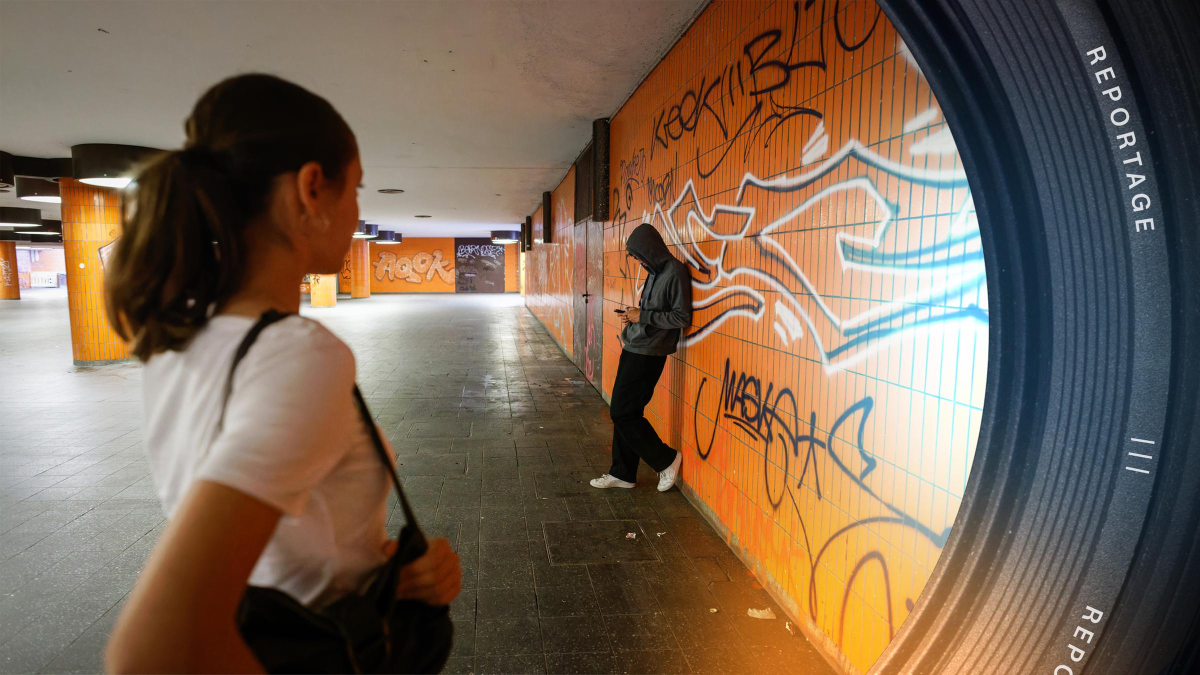 Eine junge Frau läuft durch eine Unterführung und schaut zu einen Mann im Hoodie, der an der Wand lehnt und auf sein Handy schaut. Sie hält ihre Handtasche fest.