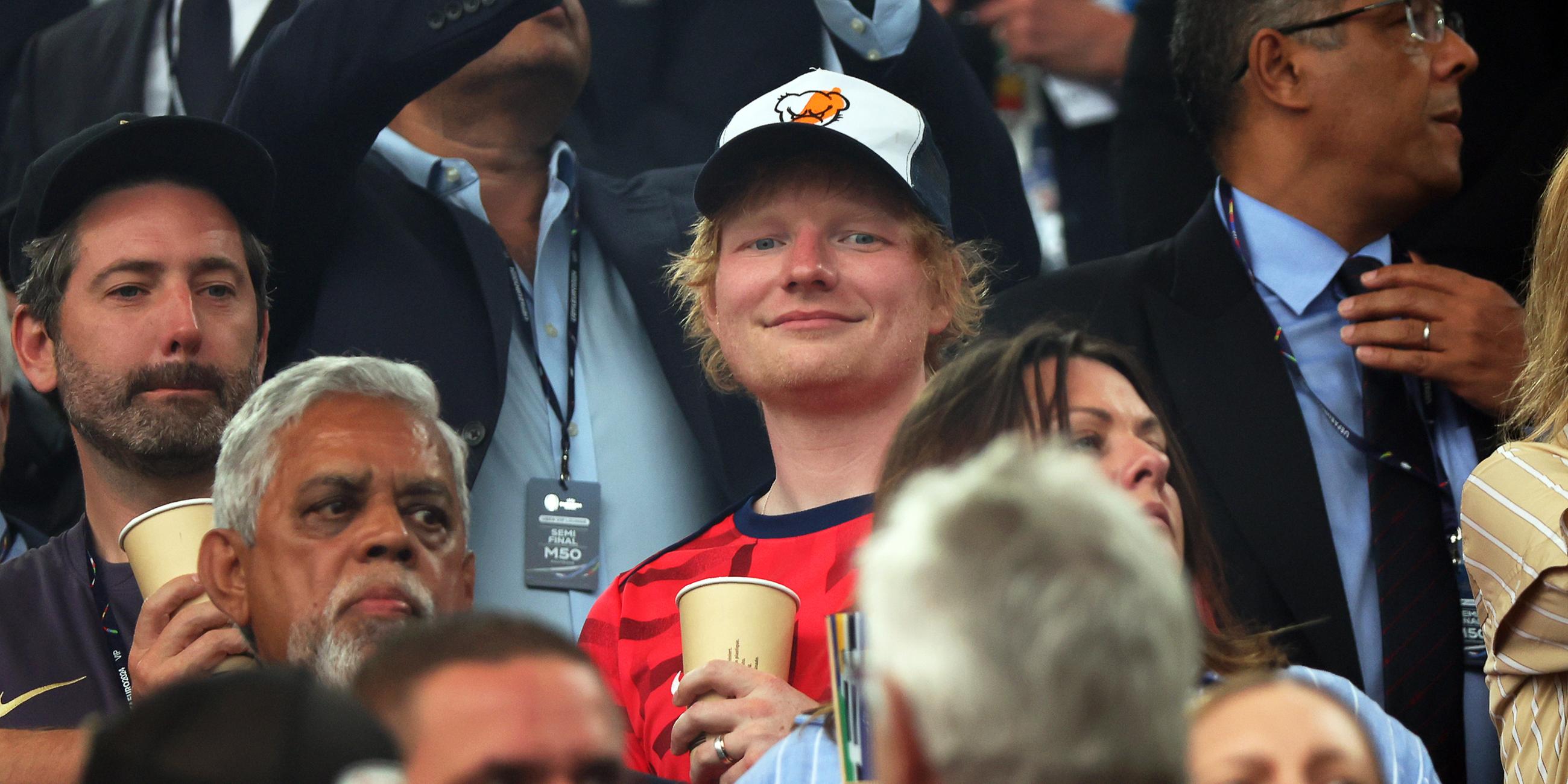 Ed Sheeran (M), Sänger aus Großbritannien, steht vor Spielbeginn auf der Tribüne.