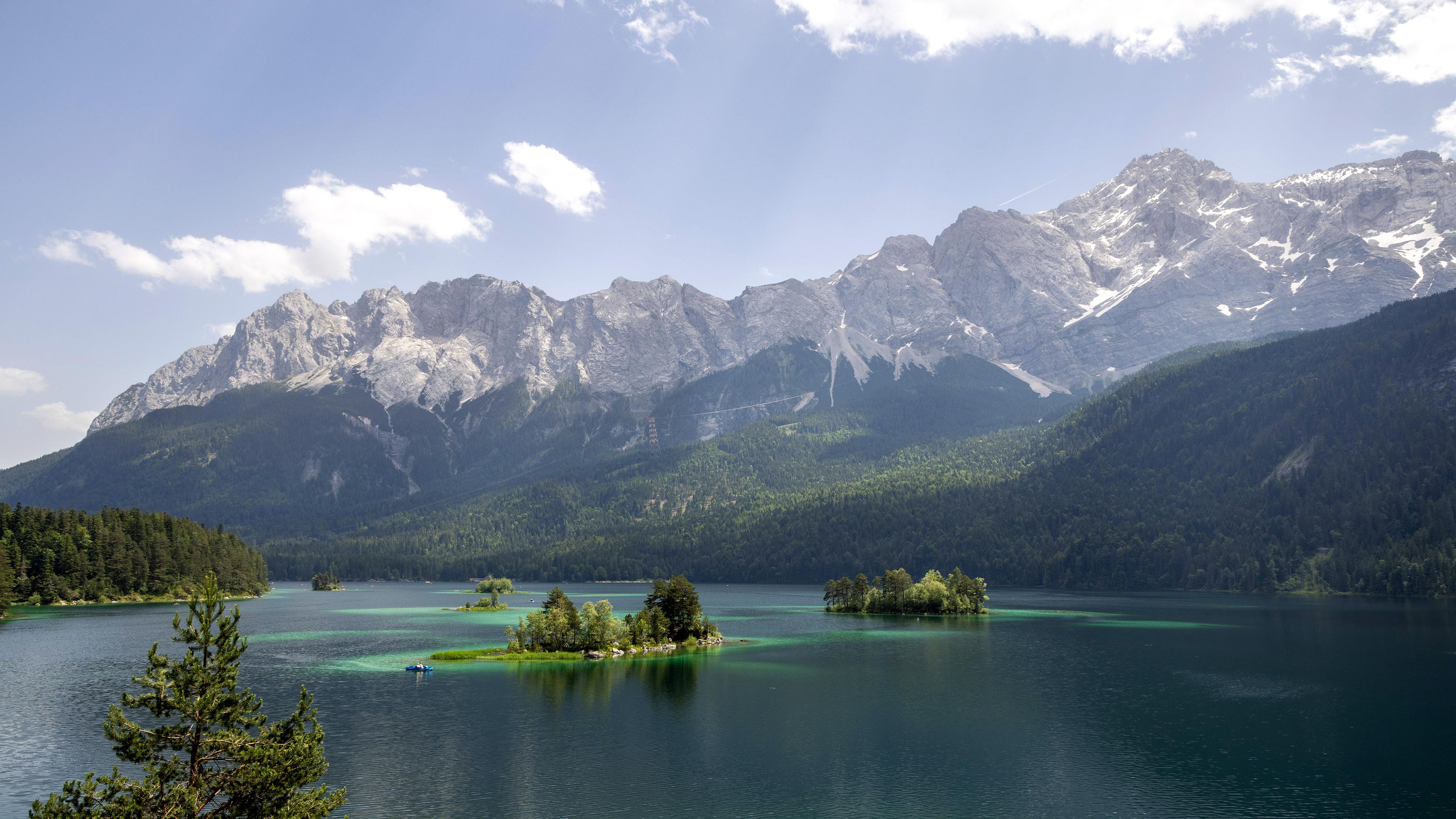 Die Sonne scheint bei blauem Himmel und wenigen Wolken auf die Landschaft am Eibsee, im Hintergrund ist das Zugspitzmassiv zu sehe