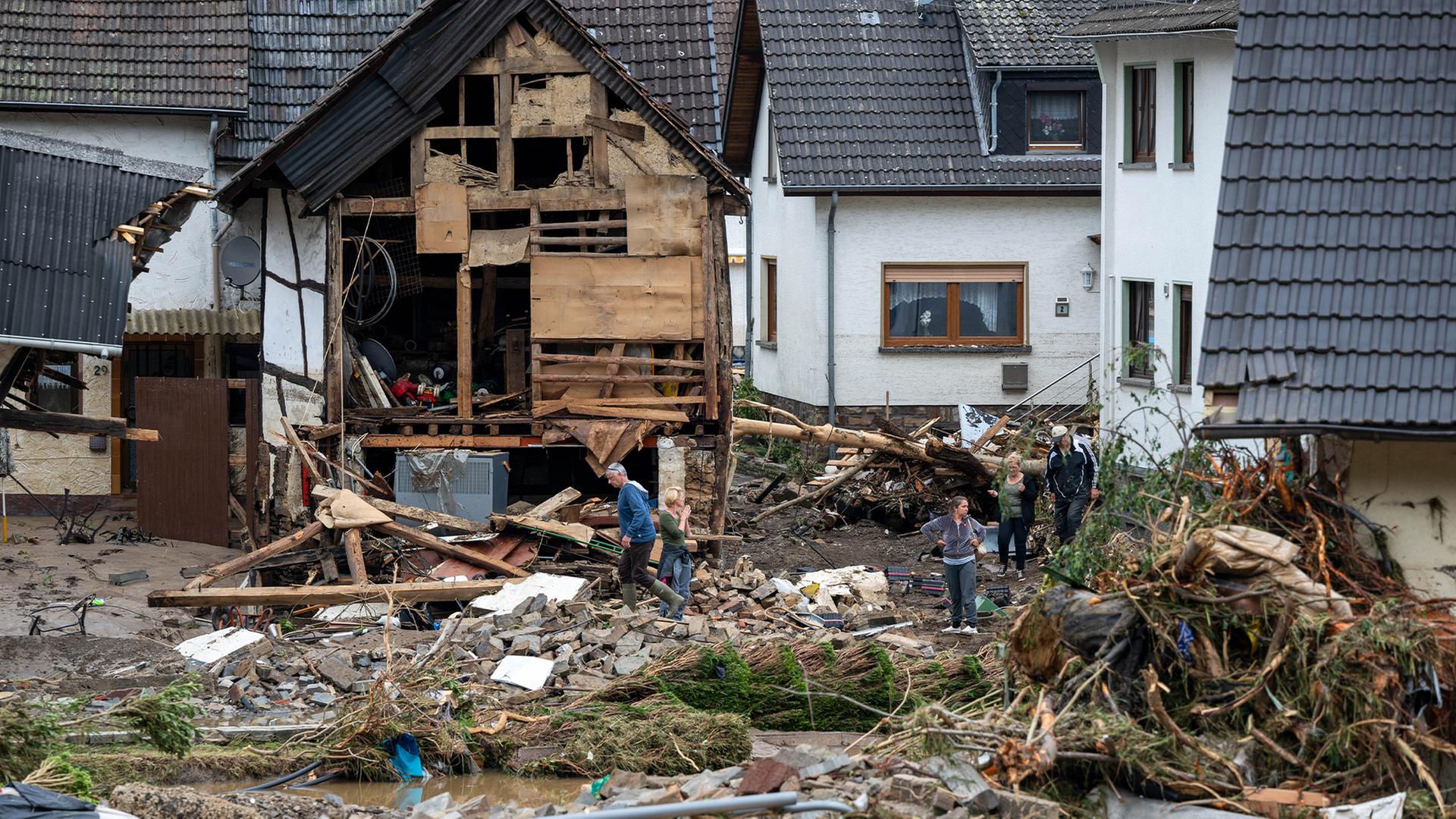 Im Hintergrund zerstörte Häuser, im Vordergrund Trümmer auf dem Boden. Dazwischen einige Menschen, die versuchen aufzuräumen.