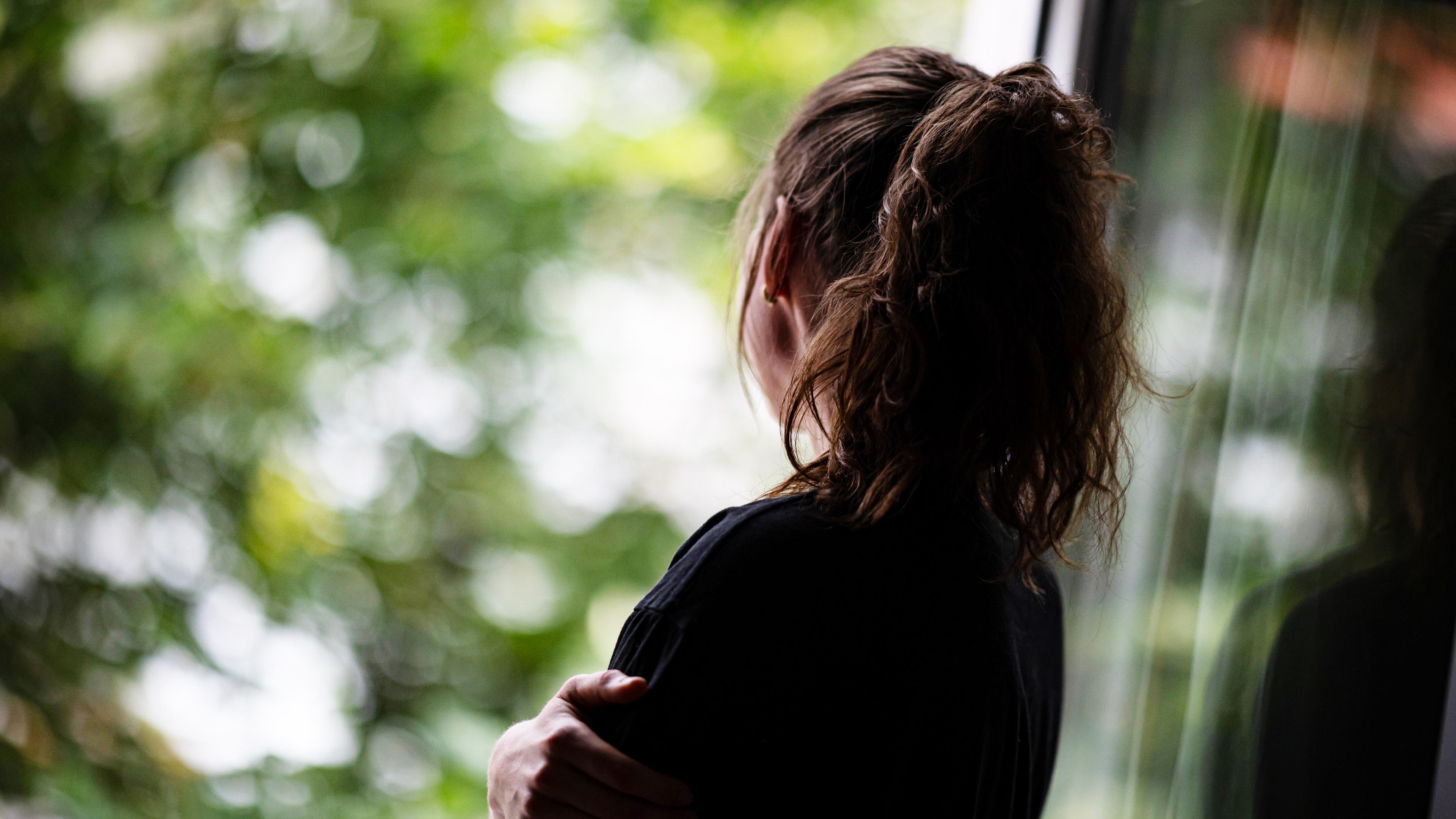 Rückansicht einer einsam wirkenden Frau, die an einem Fenster steht und hinausschaut.