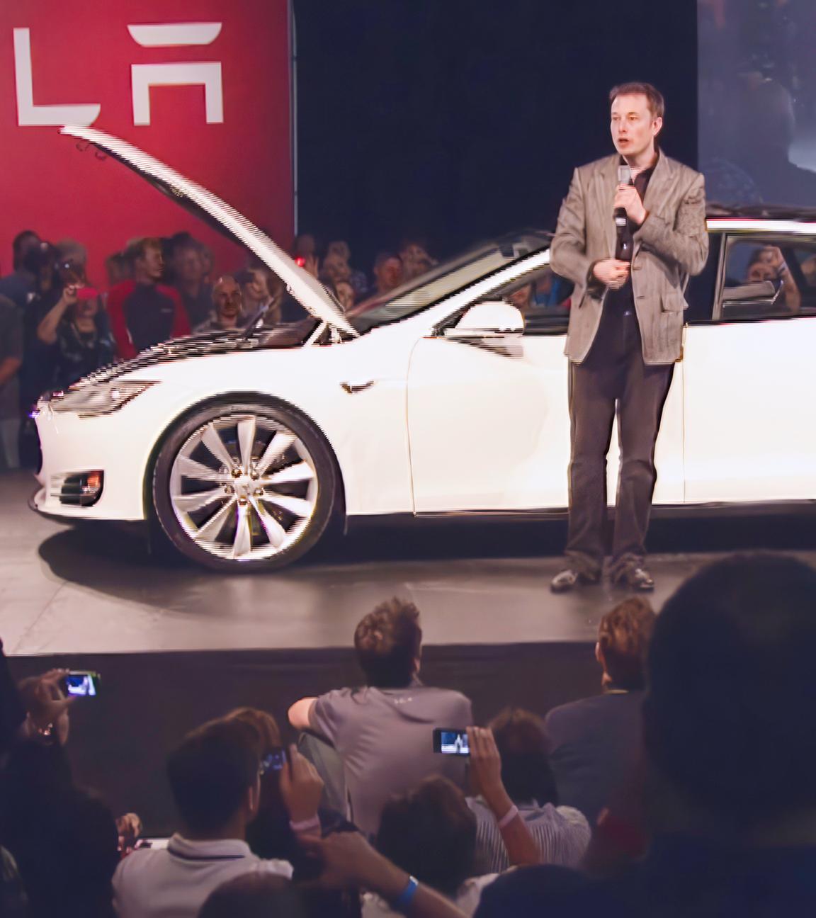 Elon Musk steht auf einer Bühne vor einem weißen Auto. Um die Bühne herum befinden sich eine Vielzahl von Menschen.