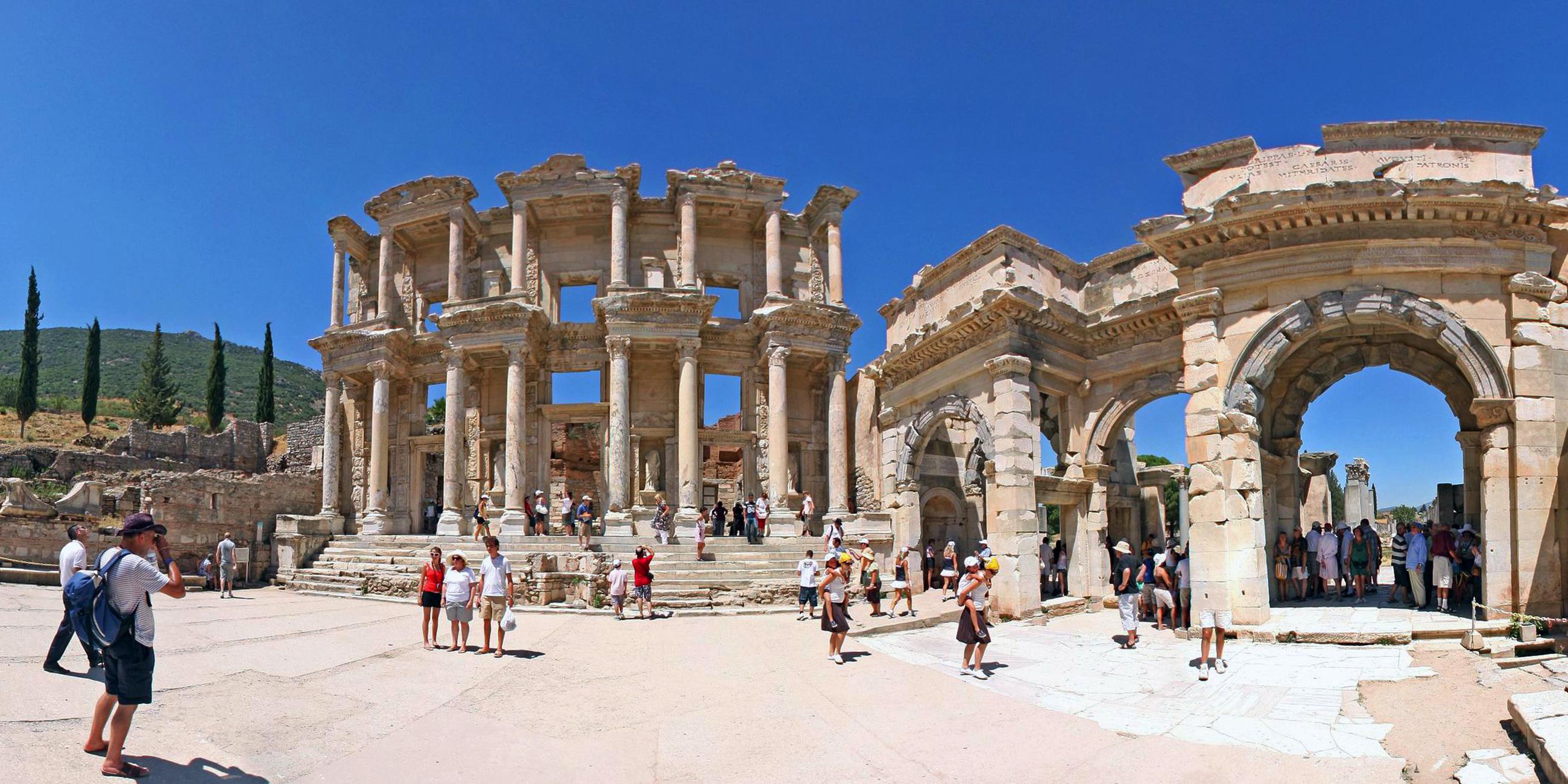 Ruinen einer antiken Bibliothek in Ephesus in der Türkei. Strahlendblauer Himmel, Touristen schauen sich die Säulen, Treppen und Fassadenteile an.