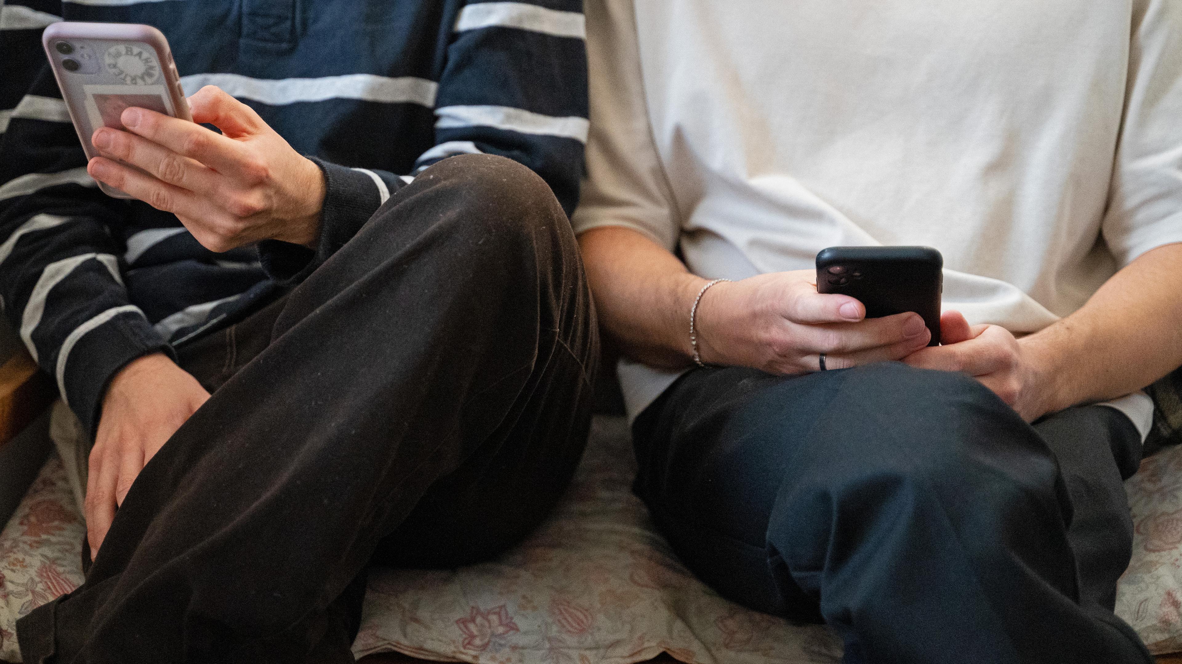 Zwei junge Männer sitzen nebeneinander auf einer Bank und halten ihre Smartphones in den Händen.