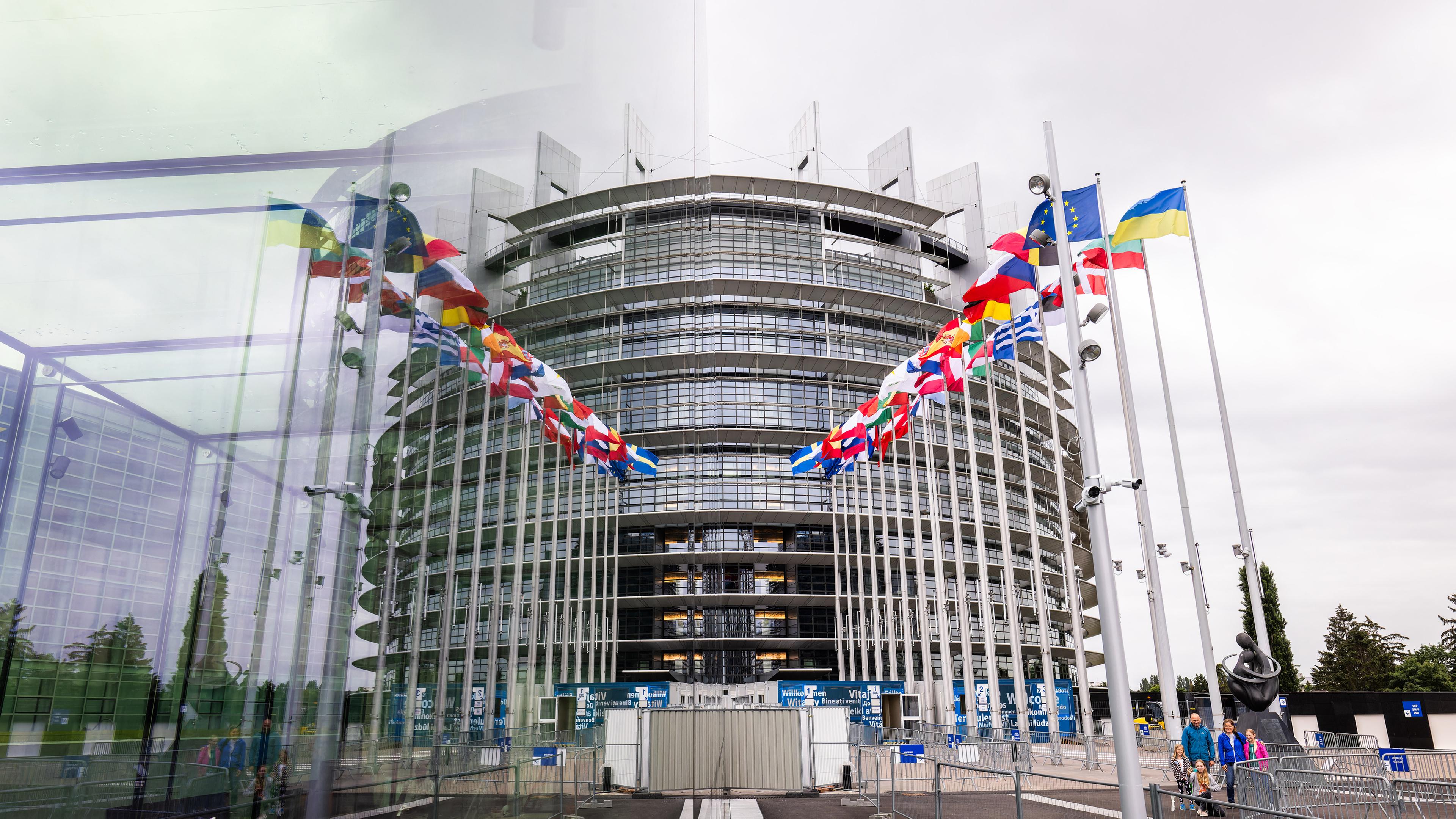Das EU Parlament ist von außen zu sehen. Vor dem runden Gebäude sind alle Nationalflaggen aufgestellt.