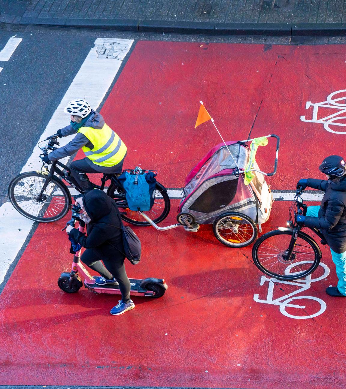 Ein Scooter-Fahrer und zwei Radfahrer mit Fahrradanhängern warten an einer Kreuzung, von oben fotografiert.