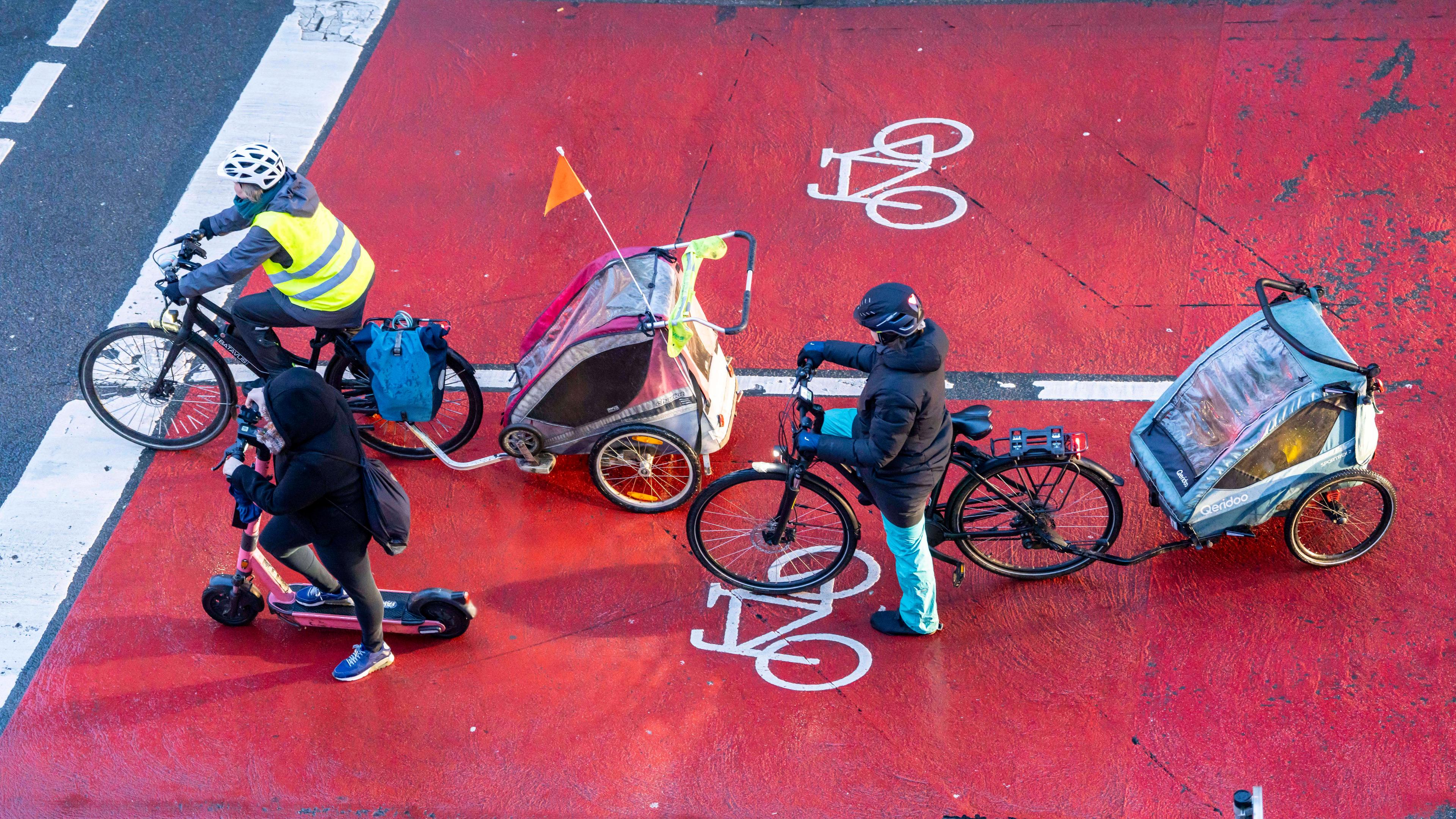 Ein Scooter-Fahrer und zwei Radfahrer mit Fahrradanhängern warten an einer Kreuzung, von oben fotografiert.