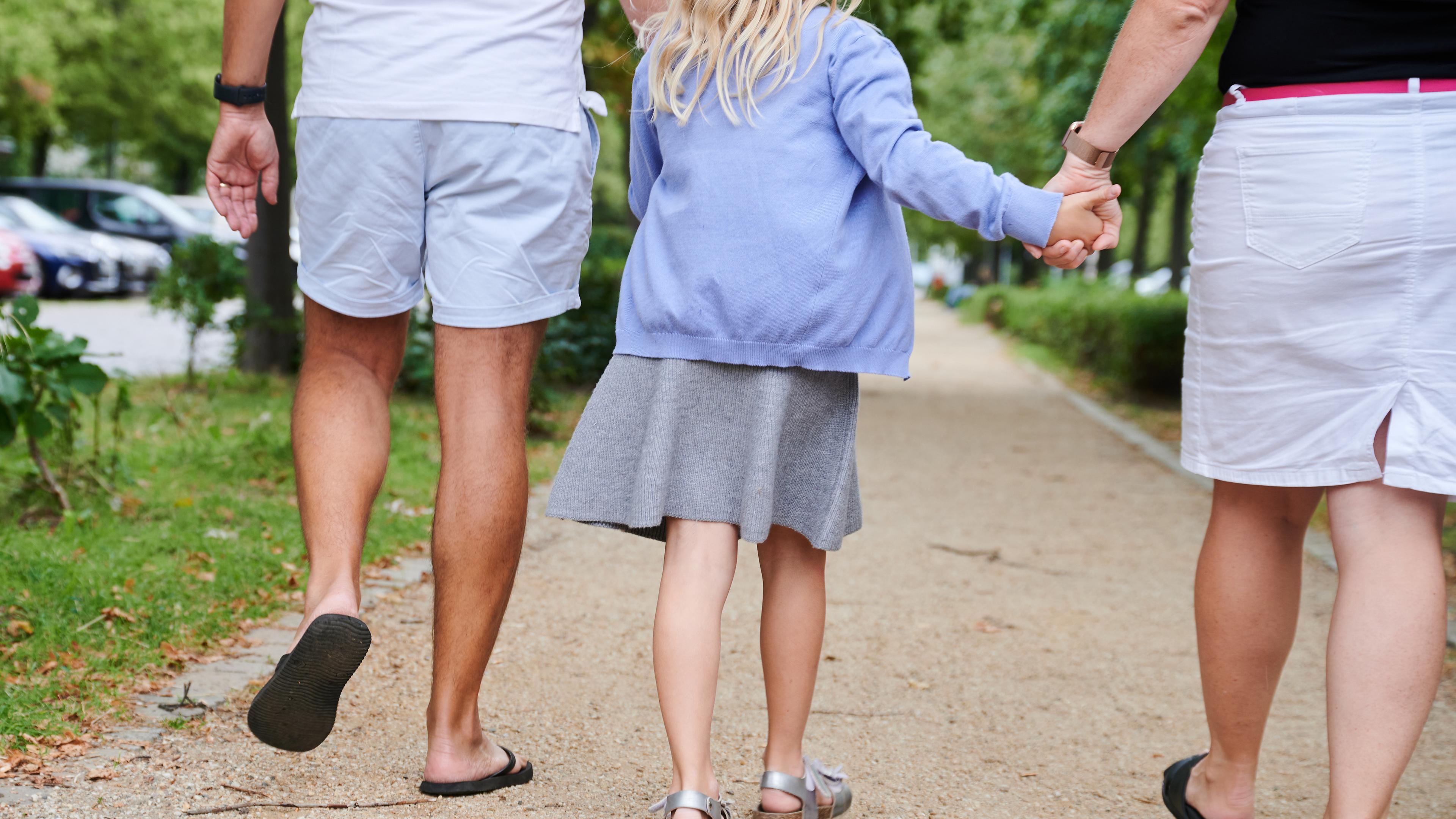 Ein Vater und eine Mutter halten die Hände ihrer Tochter, während sie auf einem Gehweg gehen. 