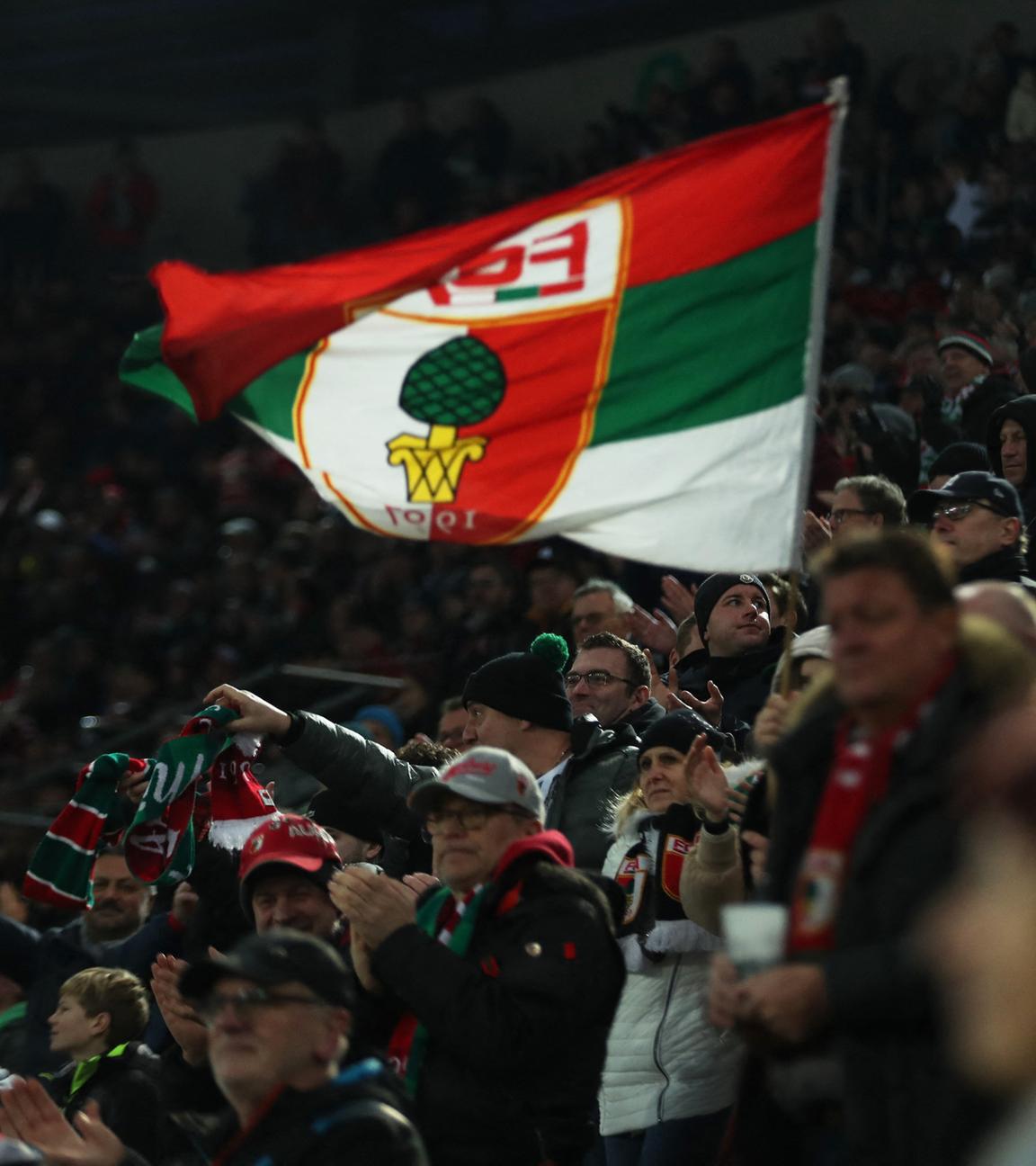 Es sind FC Augsburg Fans zu sehen, wie sie eine Flagge mit dem Vereinslogo hochhalten.