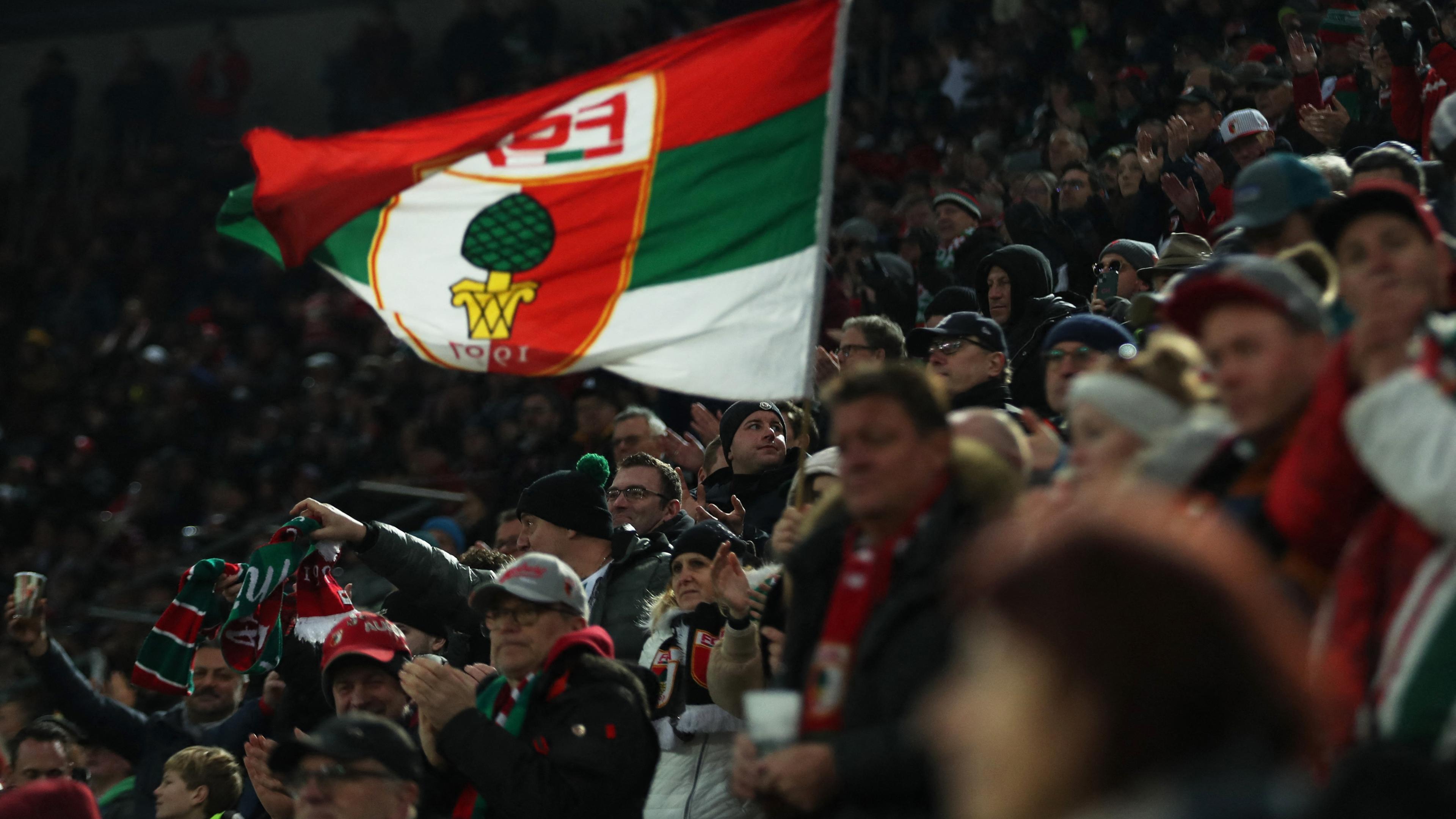 Es sind FC Augsburg Fans zu sehen, wie sie eine Flagge mit dem Vereinslogo hochhalten.