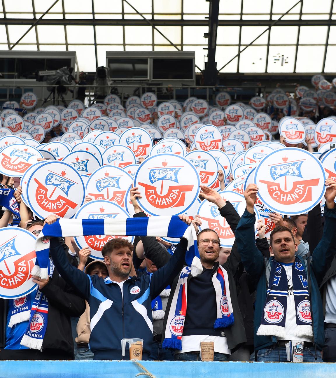 Fans vom FC Hansa Rostock stehen im Fanblock und halten Schilder mit dem Vereinslogo hoch