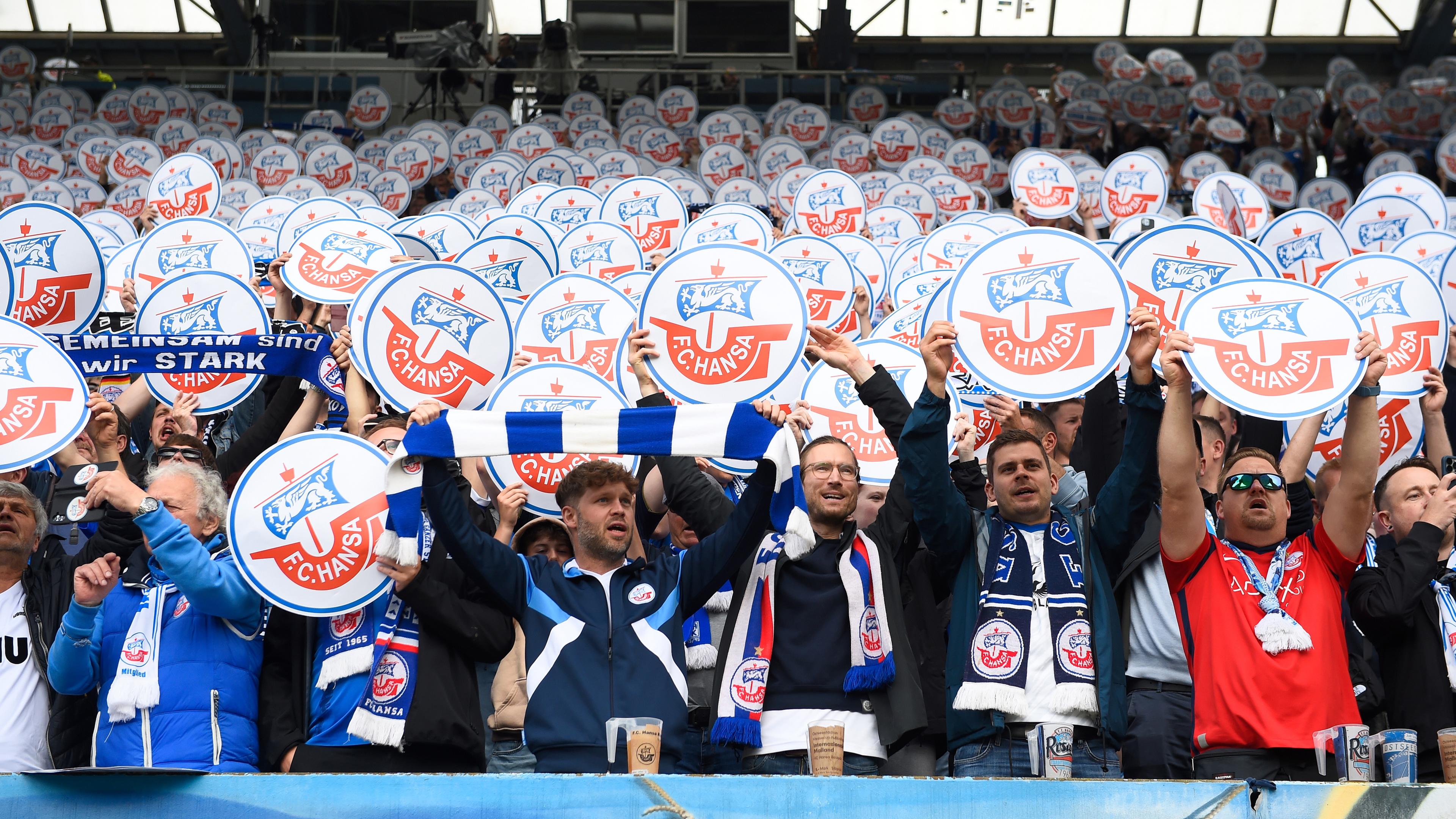 Fans vom FC Hansa Rostock stehen im Fanblock und halten Schilder mit dem Vereinslogo hoch