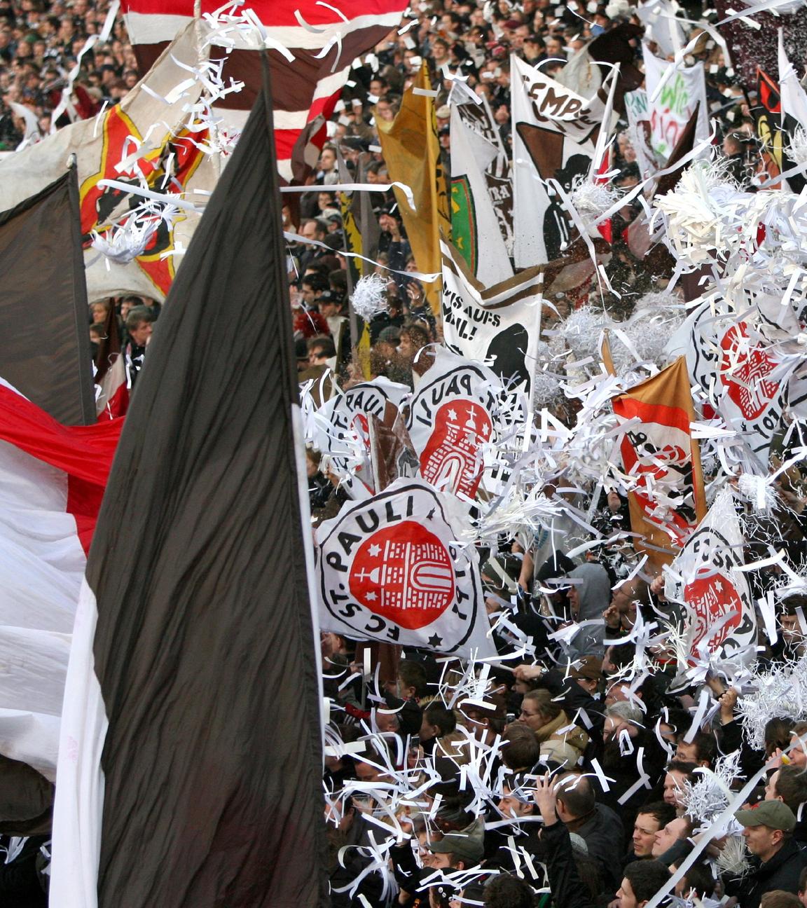 Auf einer Tribüne jubeln Fußballfans in braun, rot und weiß. Es wehen Fahnen weißes Konfetti.