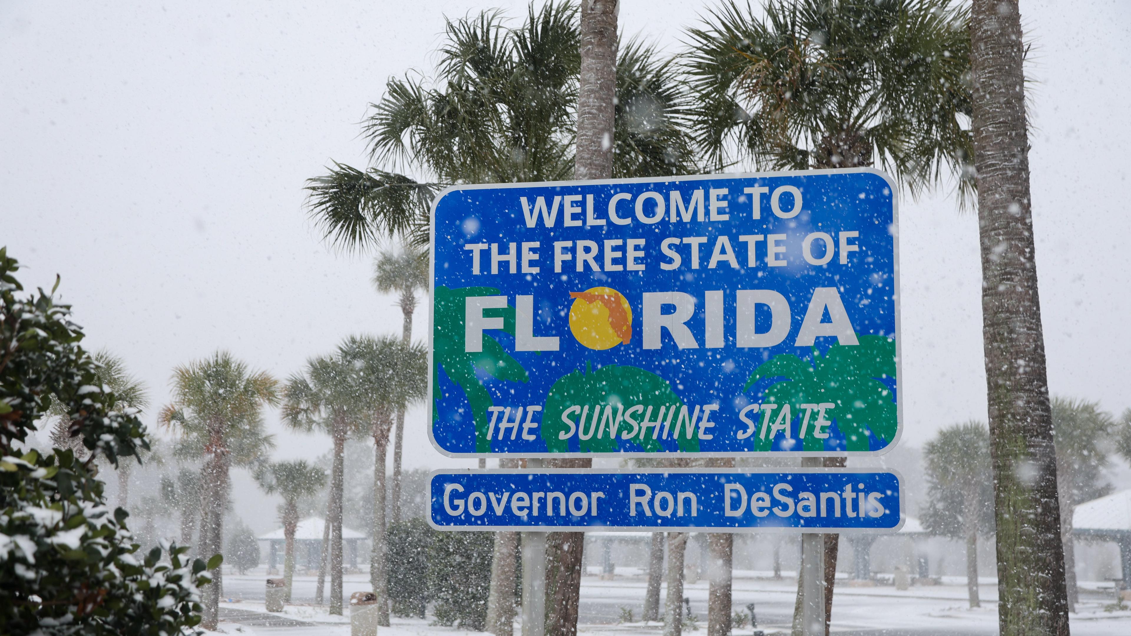 Zu sehen ist ein Staßenschild auf dem "welcome to the free state of Florida the sunshine state" steht. Rund um das Schild stehen Plamen und es liegt Schnee. Außerdem schneit es gerade.