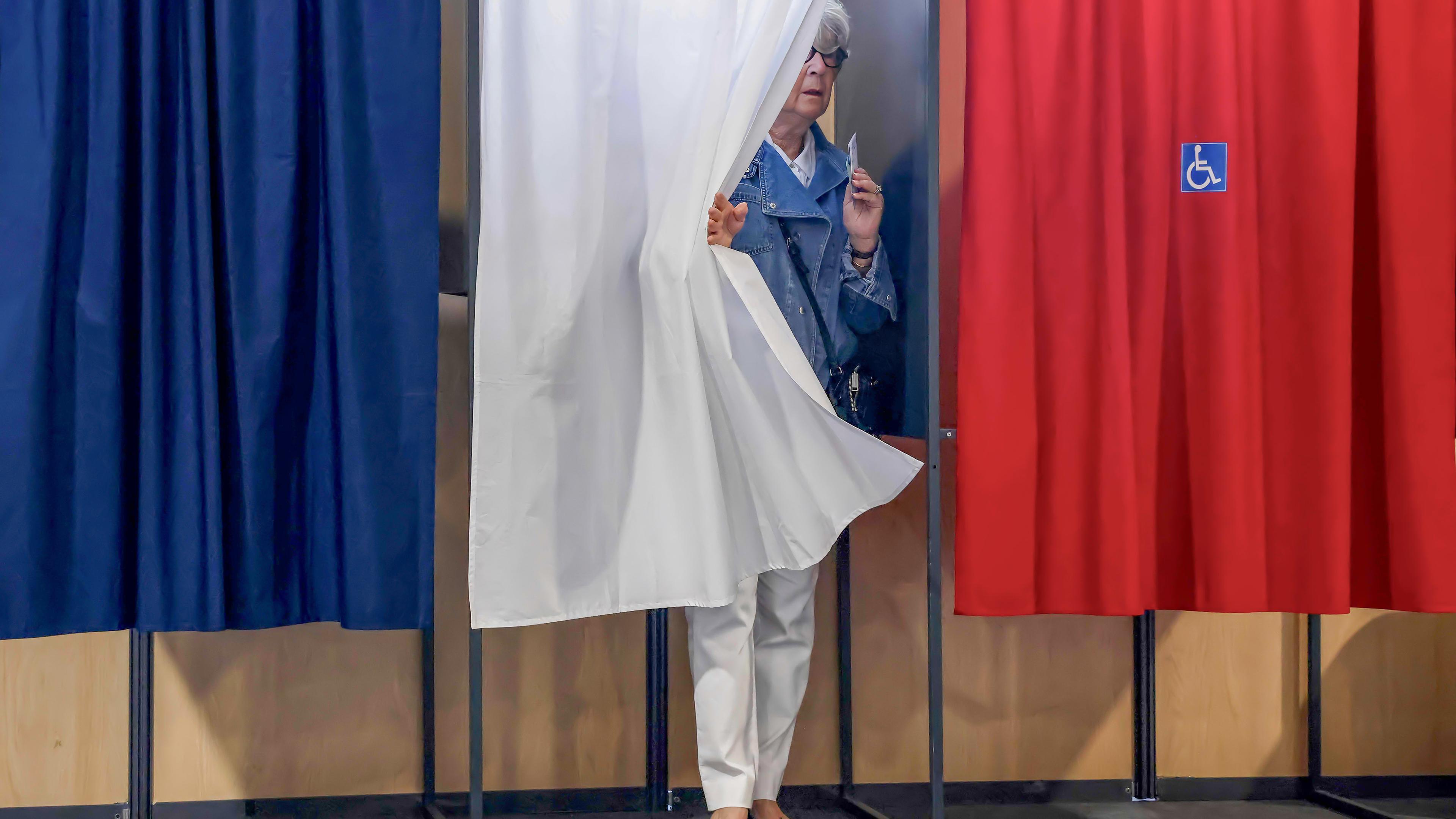 Drei Wahlkabinen sind mit einem blauen, weißen und roten Vorhang verhängt. Hinter dem mittleren weißen Vorhang tritt eine Frau mit einem Zettel in der Hand hervor.