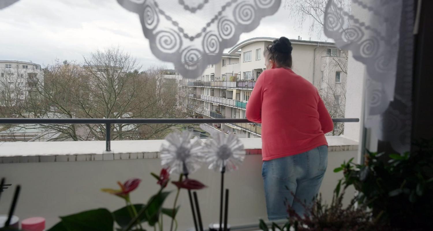 Frau steht auf dem Balkon, blickt auf Wohnsiedlung und wendet dem Betrachter den Rücken zu