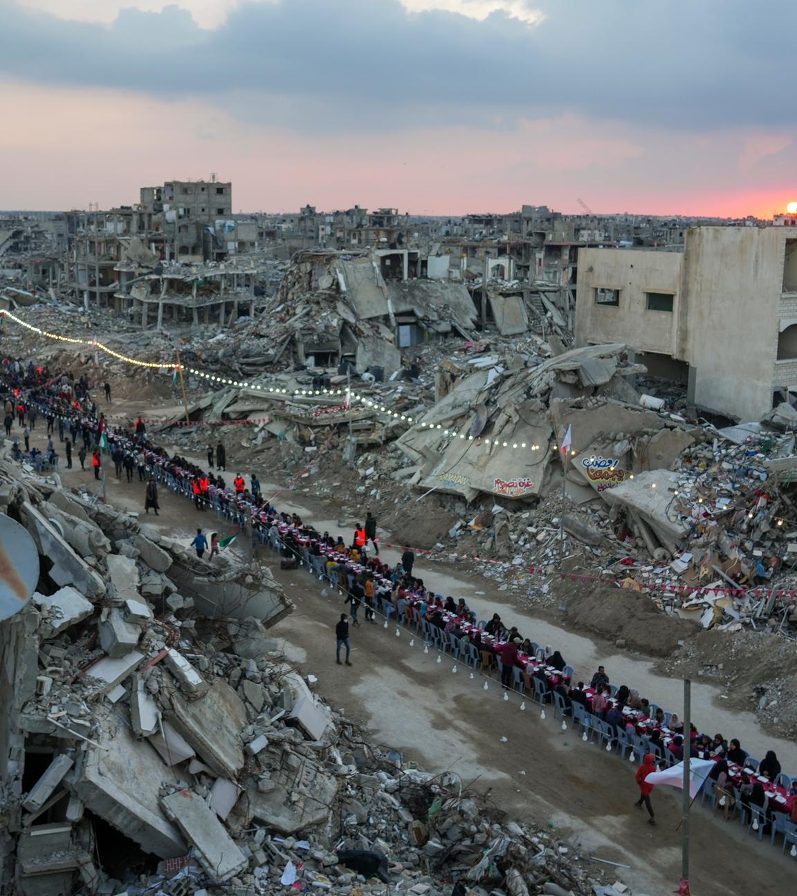 Bei Sonnenuntergang sitzen Palästinenser in Rafah an einem großen Tisch, umgeben von Trümmern zerstörter Gebäude. 