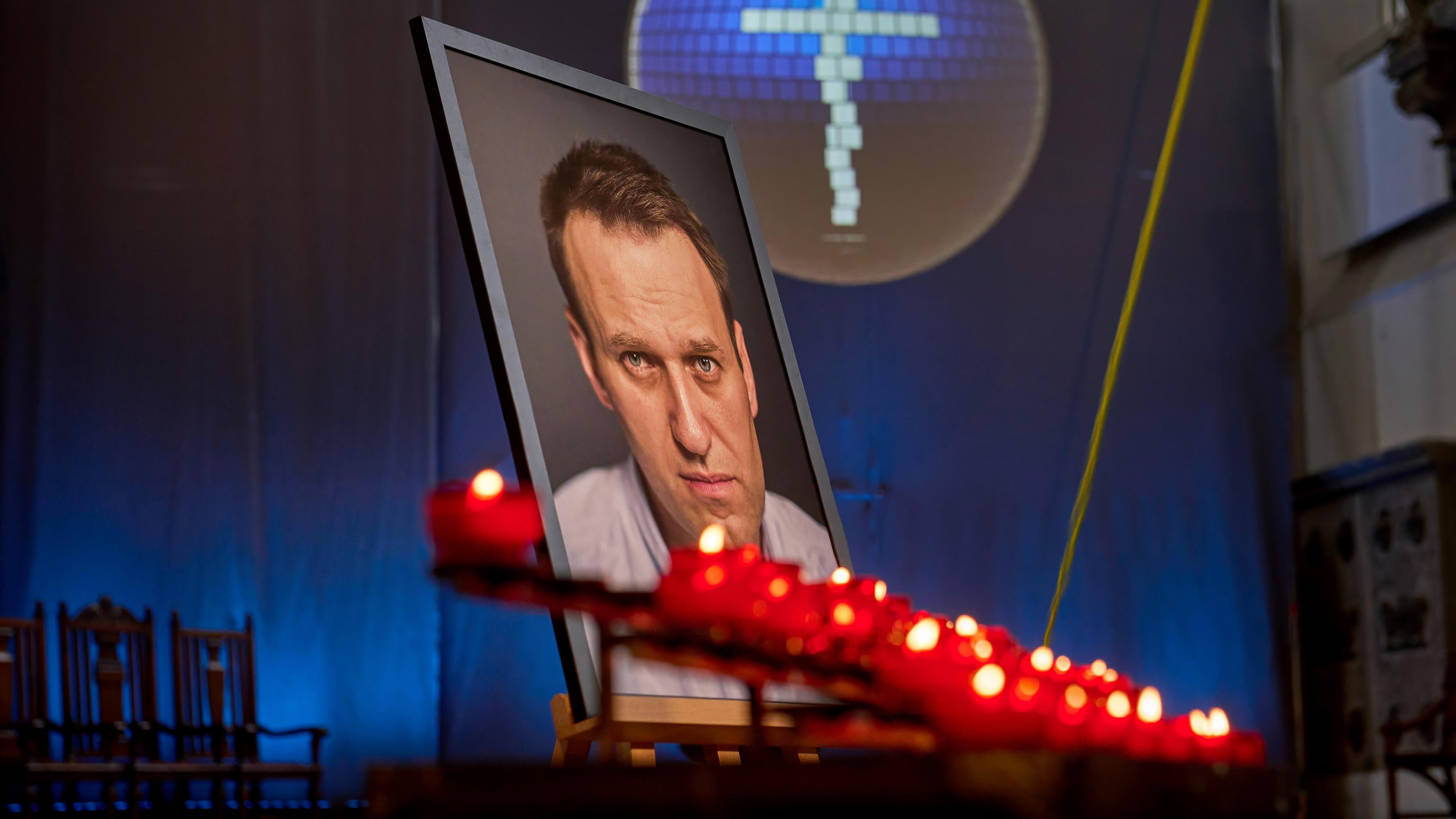 Vor dem Porträtfoto eines Mannes in einem großen Rahmen brennen Votivkerzen. Im Hintergrund ist ein Kreuz zu sehen.