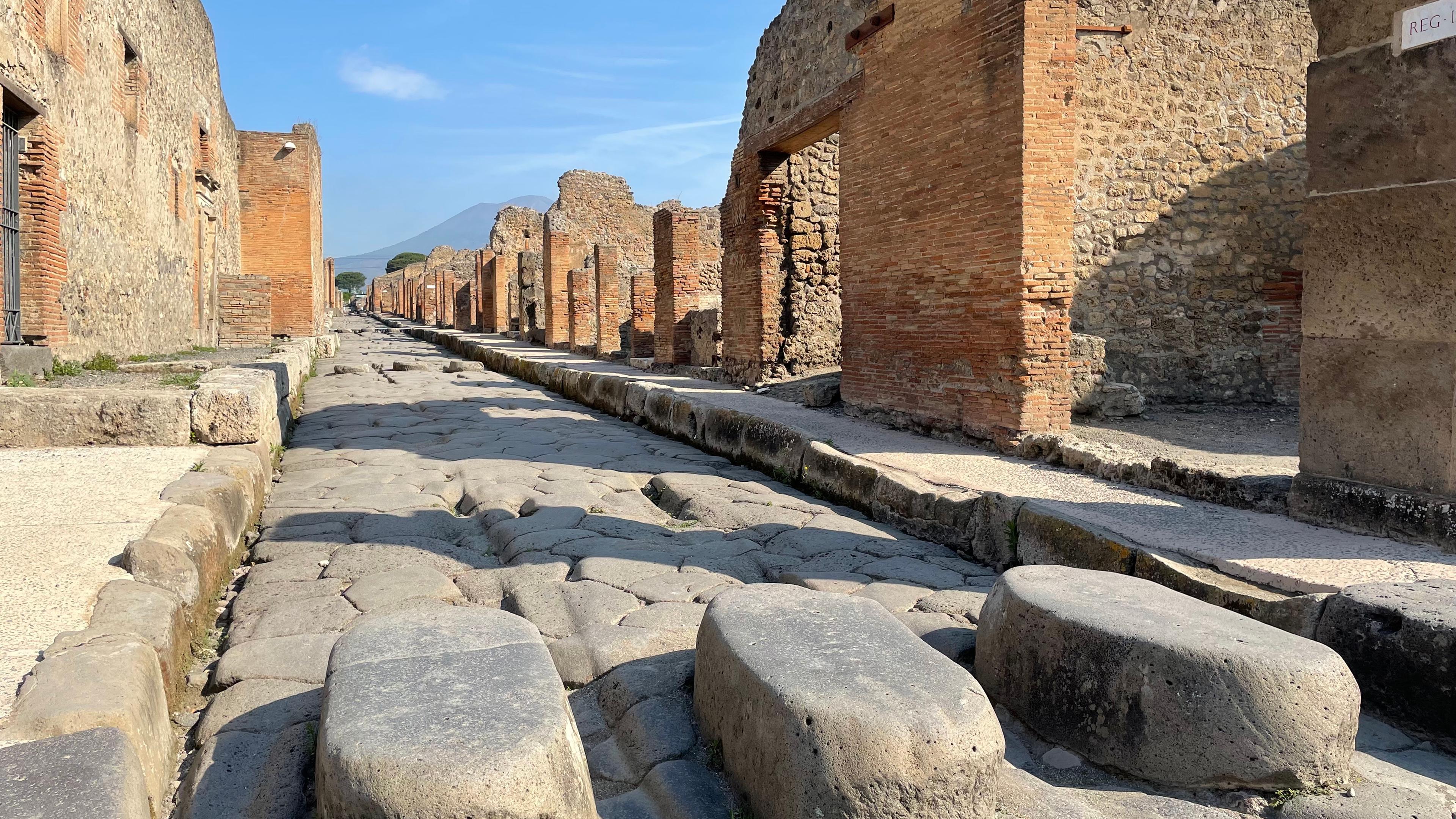 Entlang einer geraden Kopfsteinpflasterstraße im ausgegrabenen Pompeji stehen die Ruinen alter römischer Gebäude aus Ziegeln und Stein.