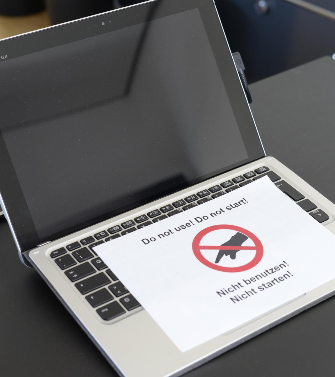 Auf einem Tisch befindet sich ein geöffneter Laptop mit schwarzem Bild. Darauf liegt ein Zettel mit dem Hinweis "Nicht benutzen!" und einer rot durchgestrichenen Hand.