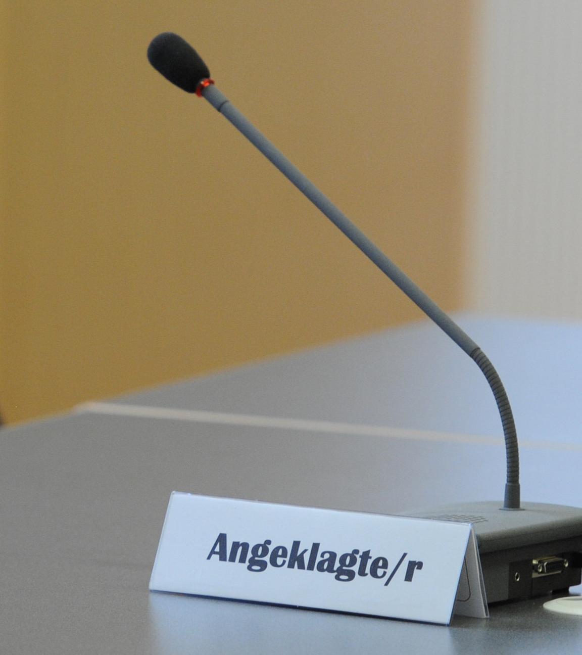 Der Sitzplatz des Angeklagten in einem Gerichtssaal. Auf dem Tisch vor dem Stuhl steht ein Mikrofon und ein Schild mit der Aufschrift "Angeklagte/r".