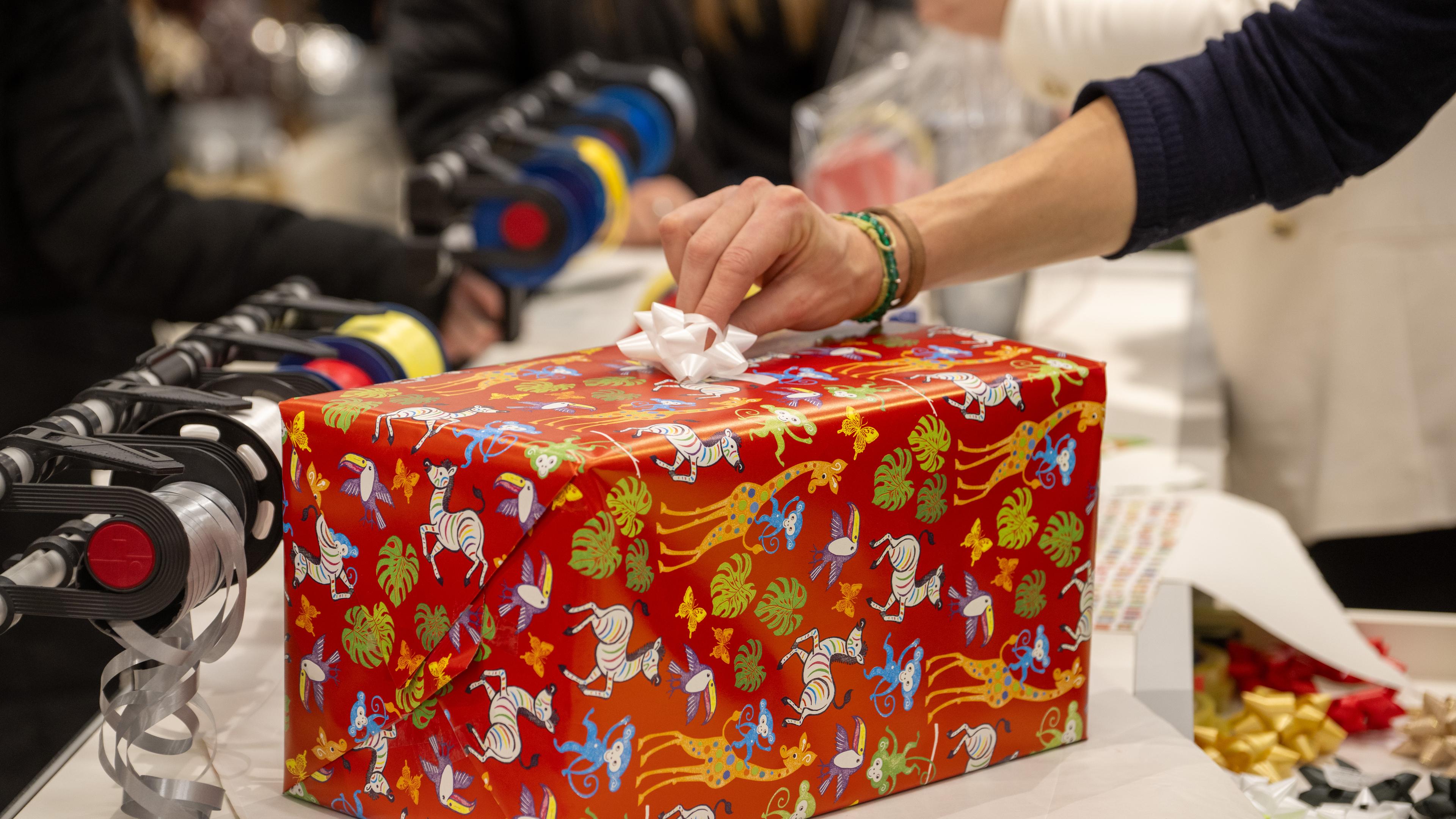 Eine Hand setzt eine Schleife auf ein rotes rechteckiges Geschenk mit Tiermuster-Geschenkpapier.