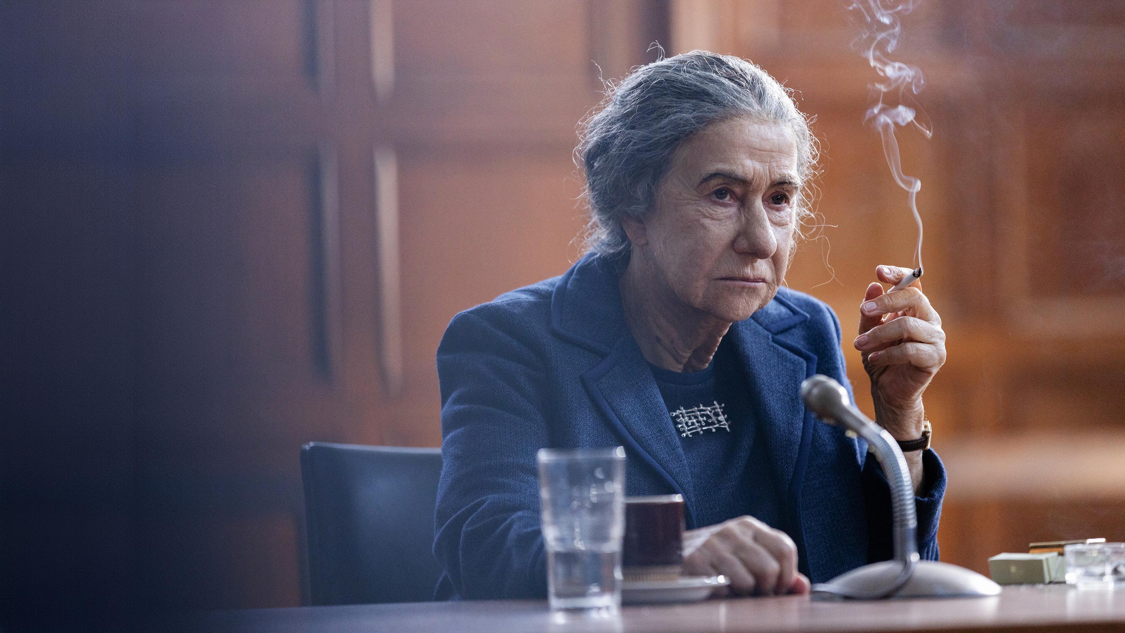 Eine ältere Frau mit zurückgebundenen Haaren sitzt rauchend und mit einer Tasse und einem Glas vor sich an einem Mikrofon.