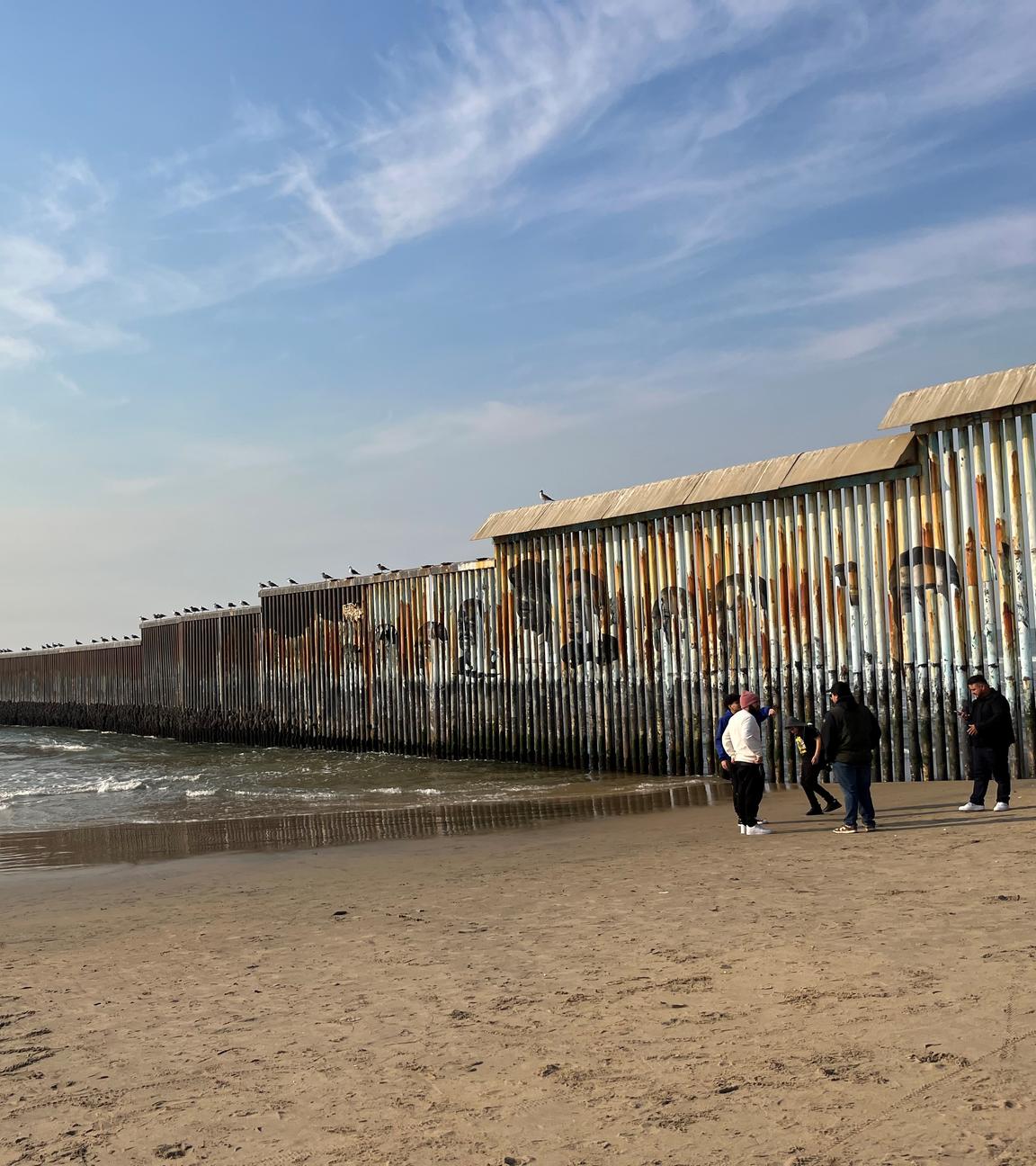 Im Hintergrund ist Meer zu sehen, vier Menschen stehen zusammen auf Sand neben einer Mauer.