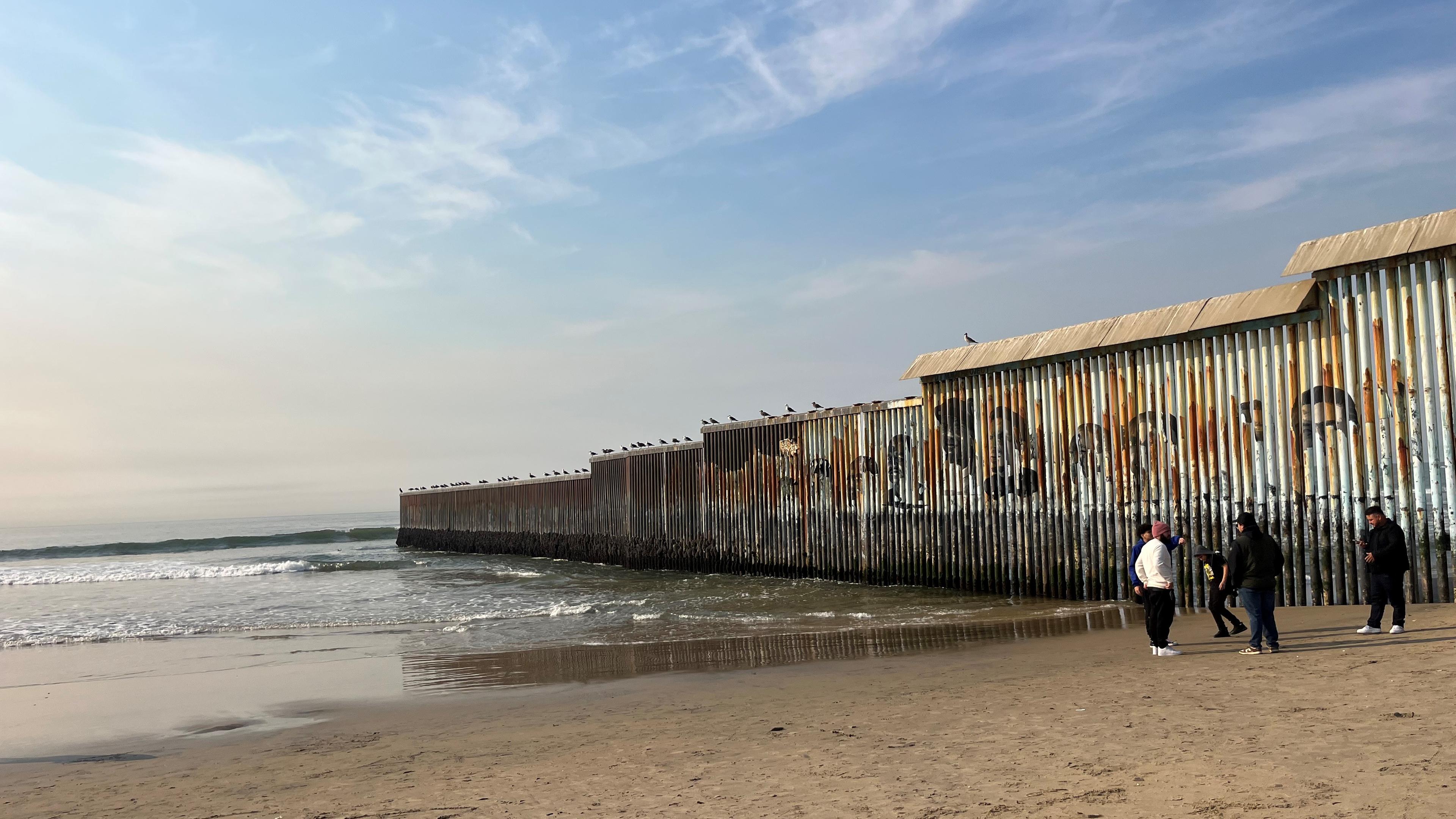 Im Hintergrund ist Meer zu sehen, vier Menschen stehen zusammen auf Sand neben einer Mauer.