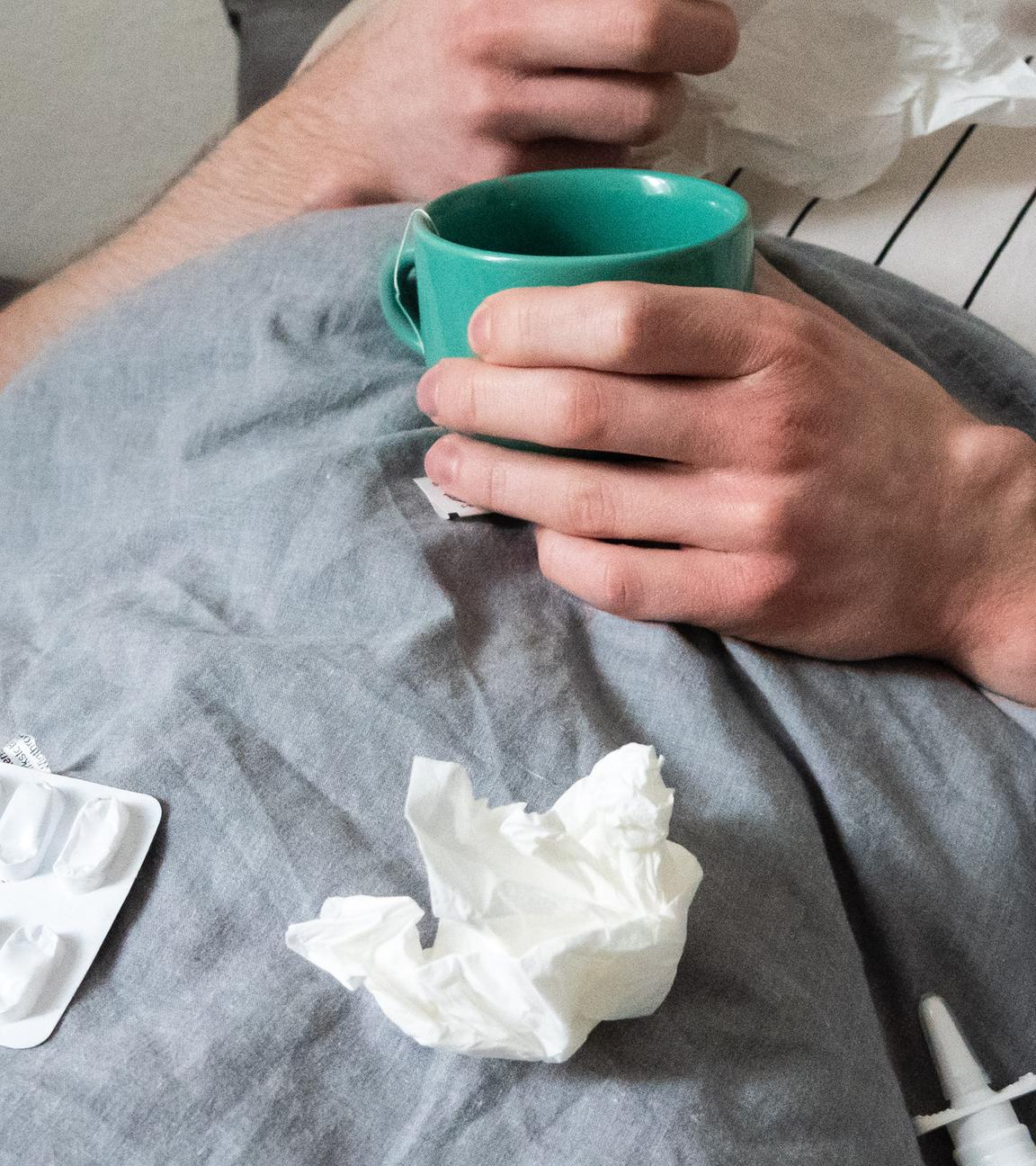 Ein Mann liegt mit Taschentüchern, Teetasse, Nasenspray und Tabletten im Bett (gestellte Szene). 