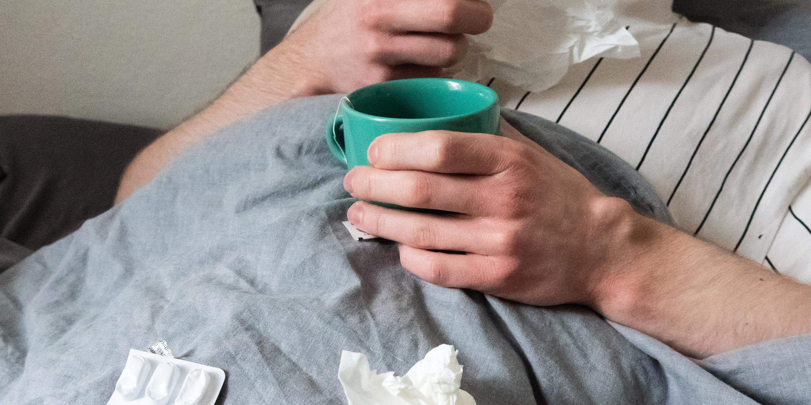 Ein Mann liegt mit Taschentüchern, Teetasse, Nasenspray und Tabletten im Bett (gestellte Szene). 