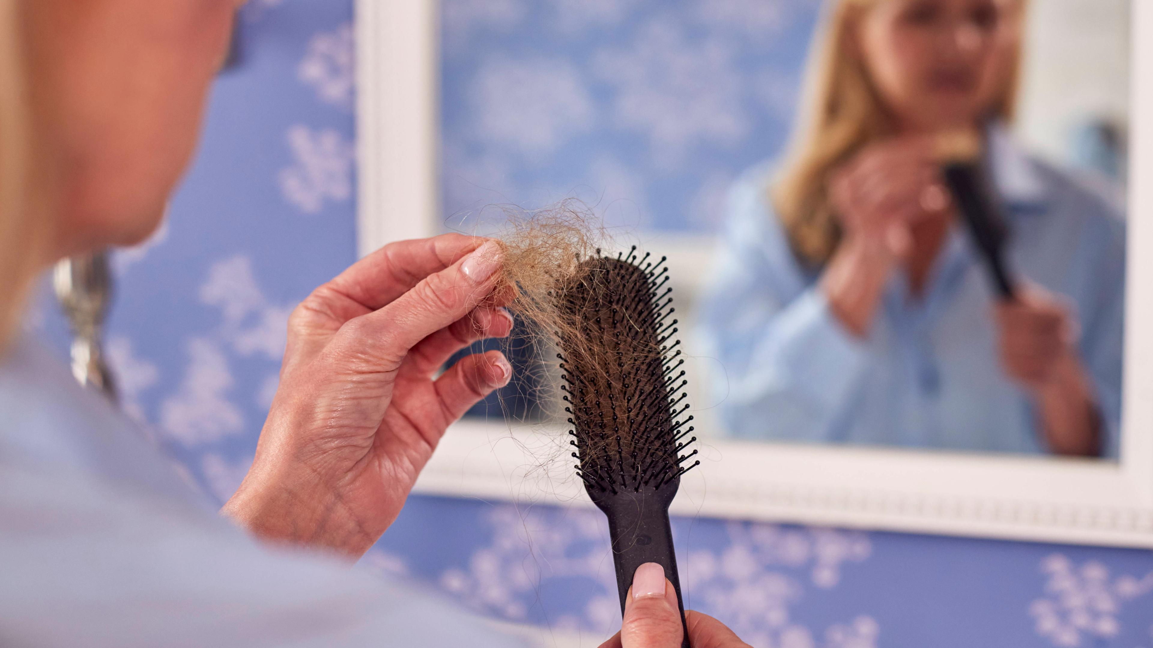 Une femme aux cheveux blonds se tient devant un miroir. Dans sa main, elle tient une brosse à cheveux dans laquelle est coincée une touffe de cheveux.