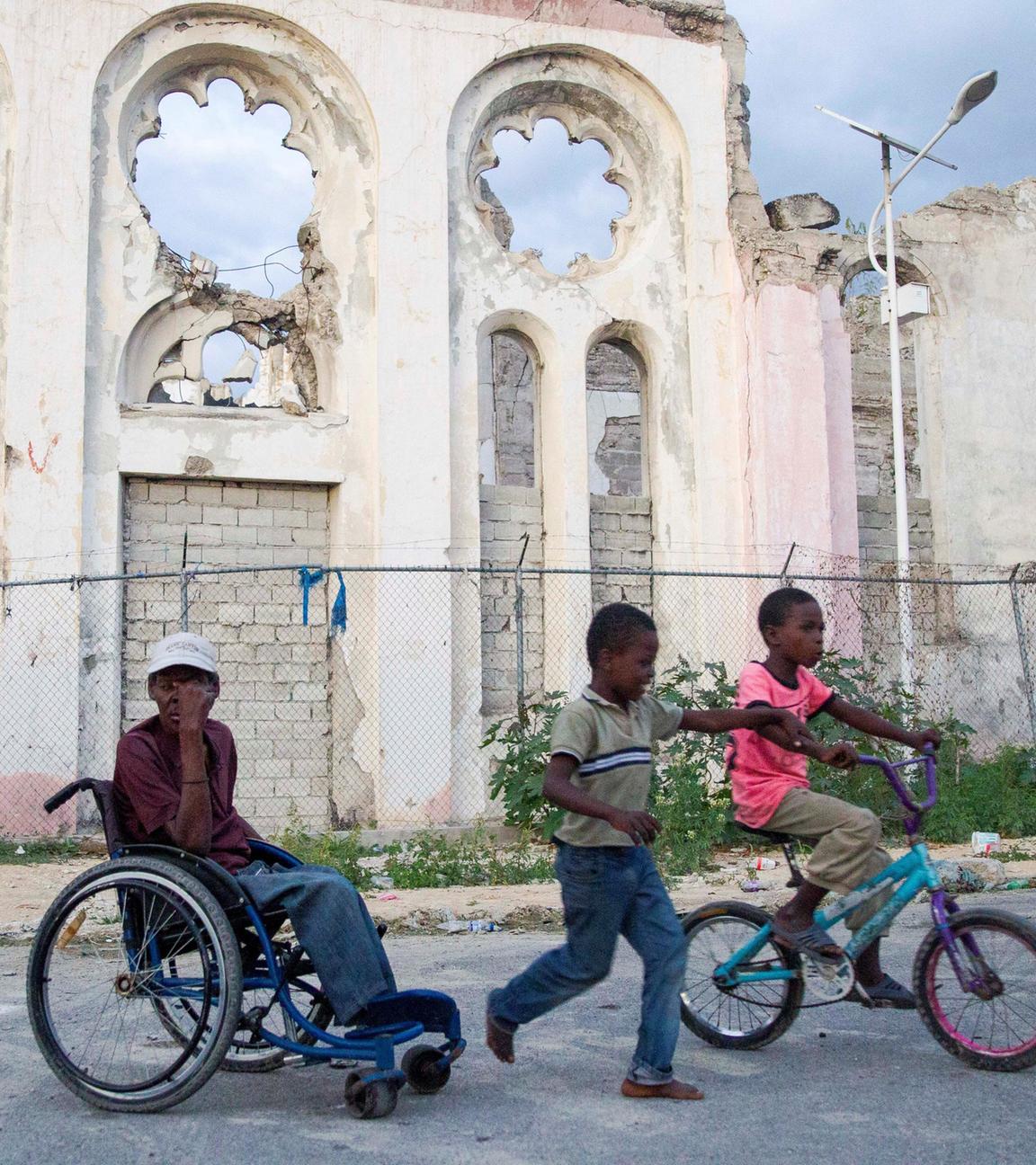 Ein Mann in einem Rollstuhl und zwei Kinder neben ihm stehen vor einer zerstörten Kirche in Port-au-Prince, Haiti.