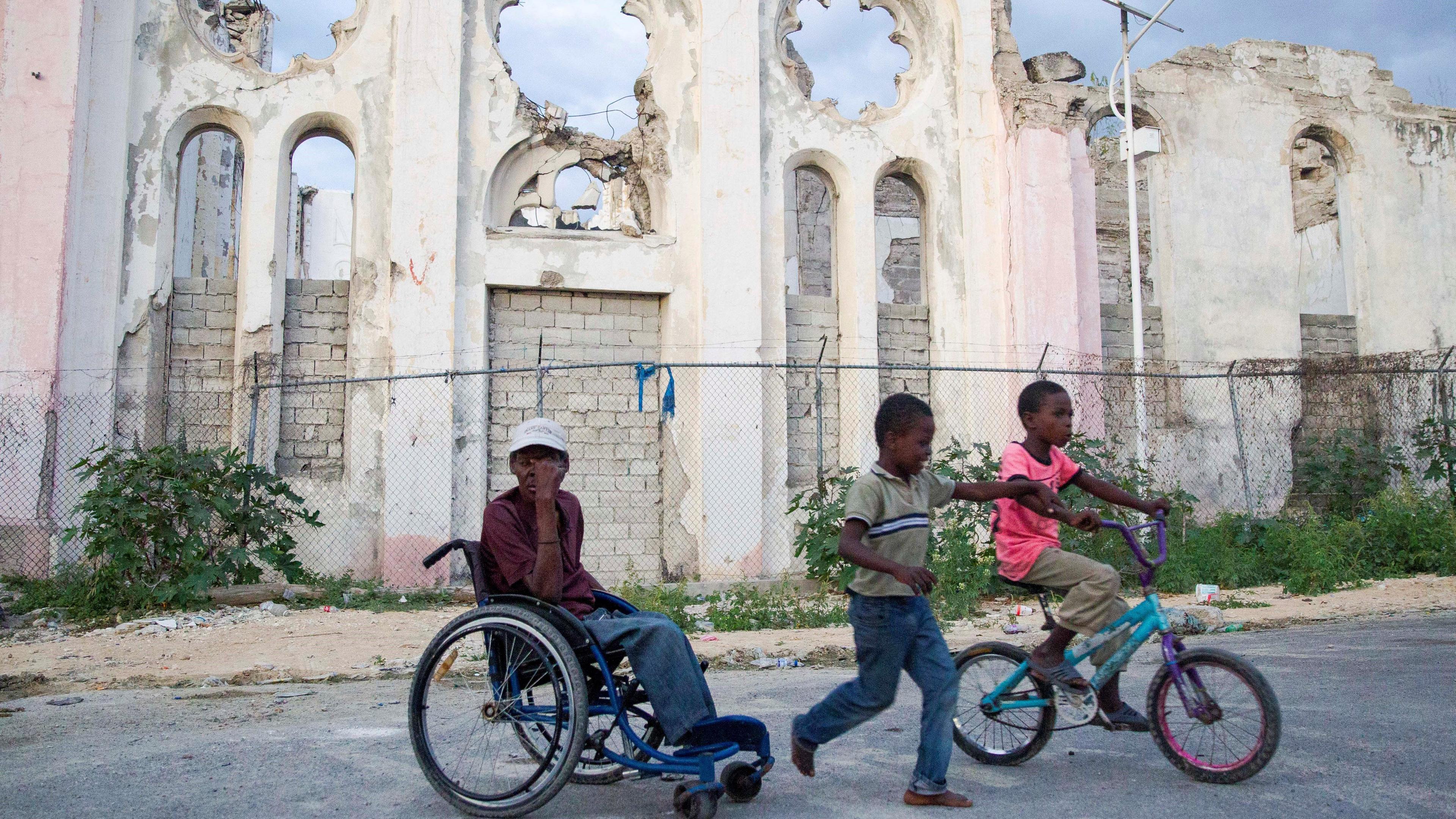 Ein Mann in einem Rollstuhl und zwei Kinder neben ihm stehen vor einer zerstörten Kirche in Port-au-Prince, Haiti.