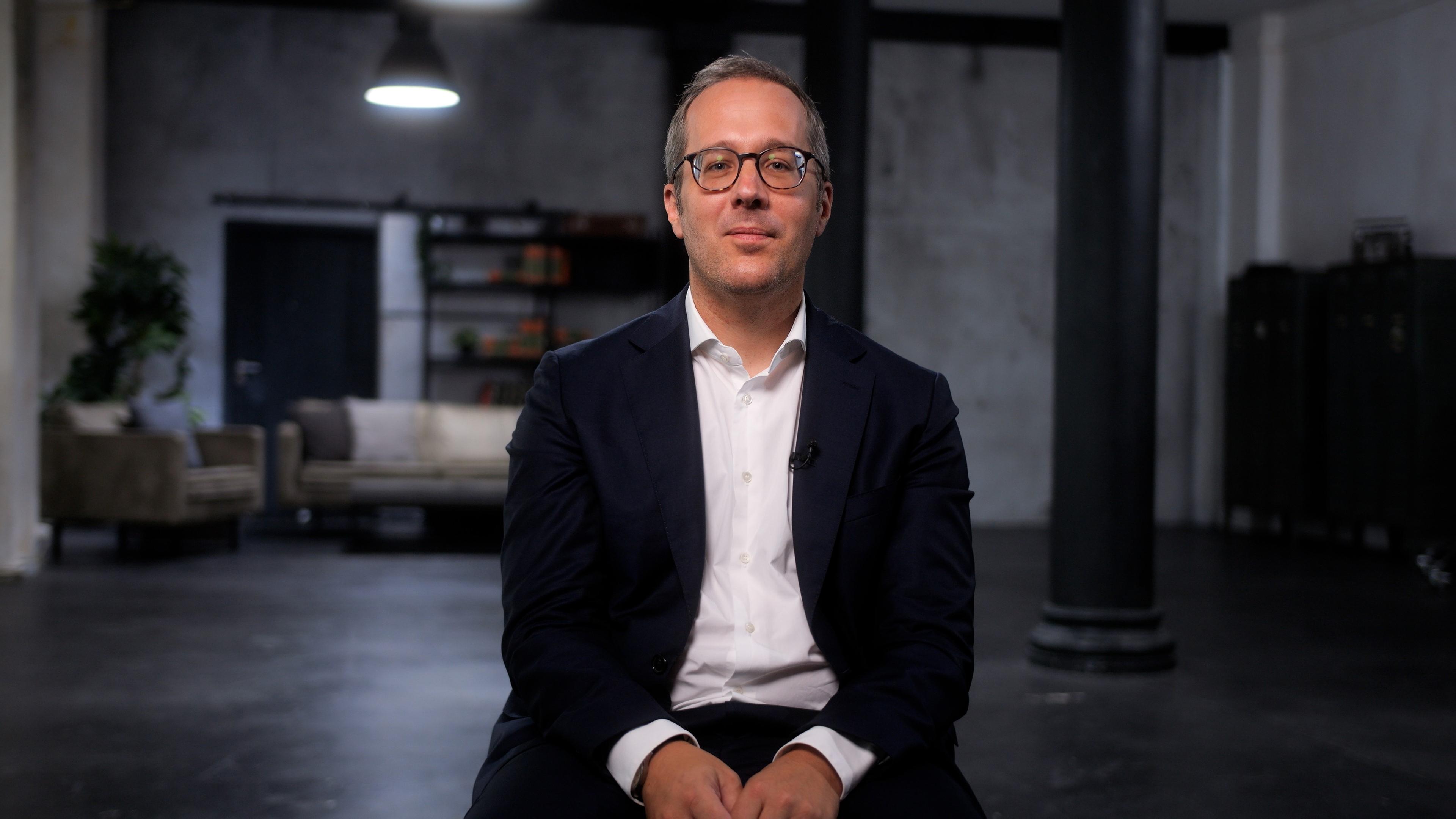 Ein mittelalter Mann mit Brille, einem weißen Hemd und Jacket sitzt.