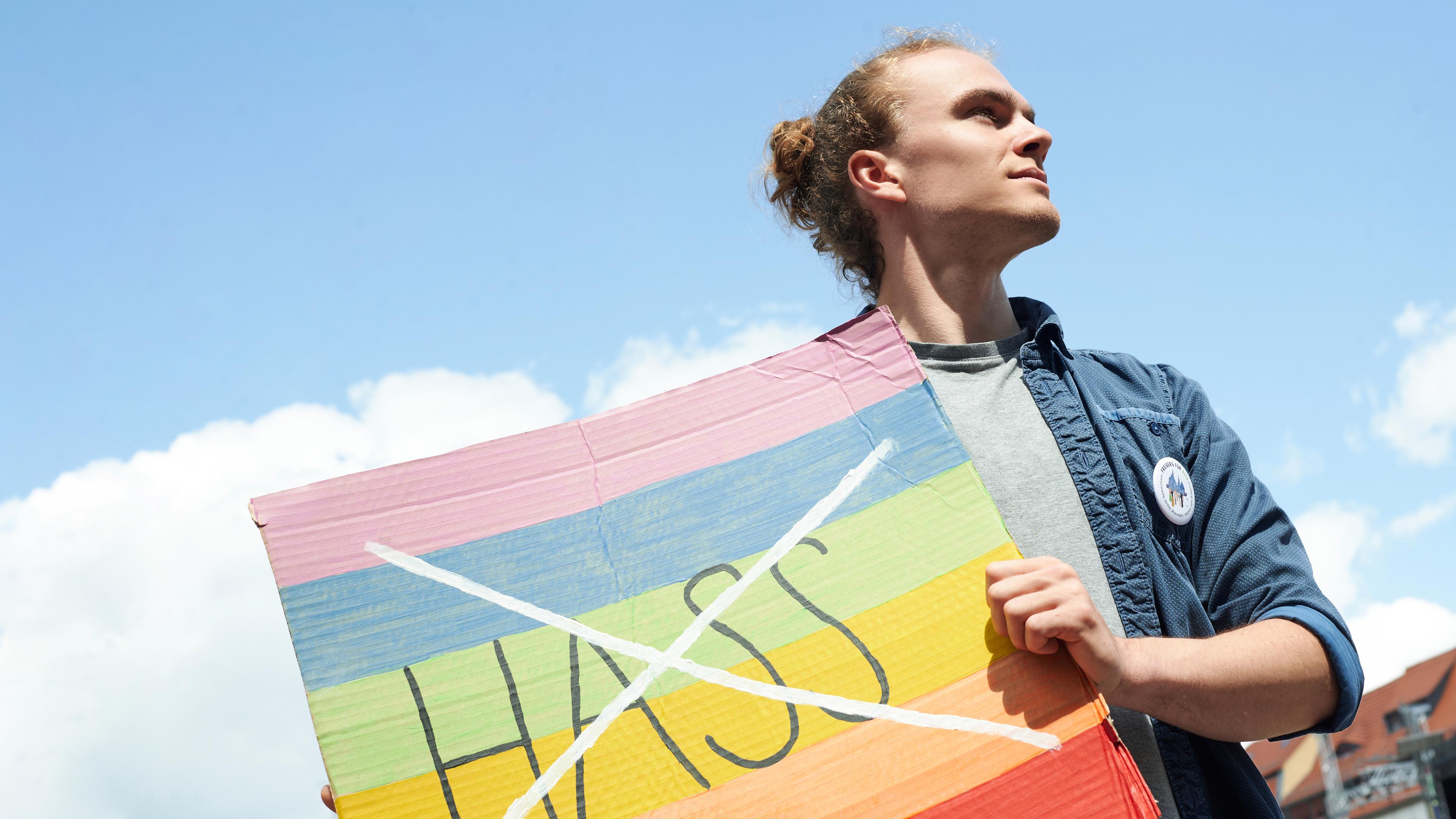 Florian hält ein buntes Schild vor seinen Körper. Auf dem Schild ist das Wort "Hass" durchgestrichen. Er blickt zur Seite.