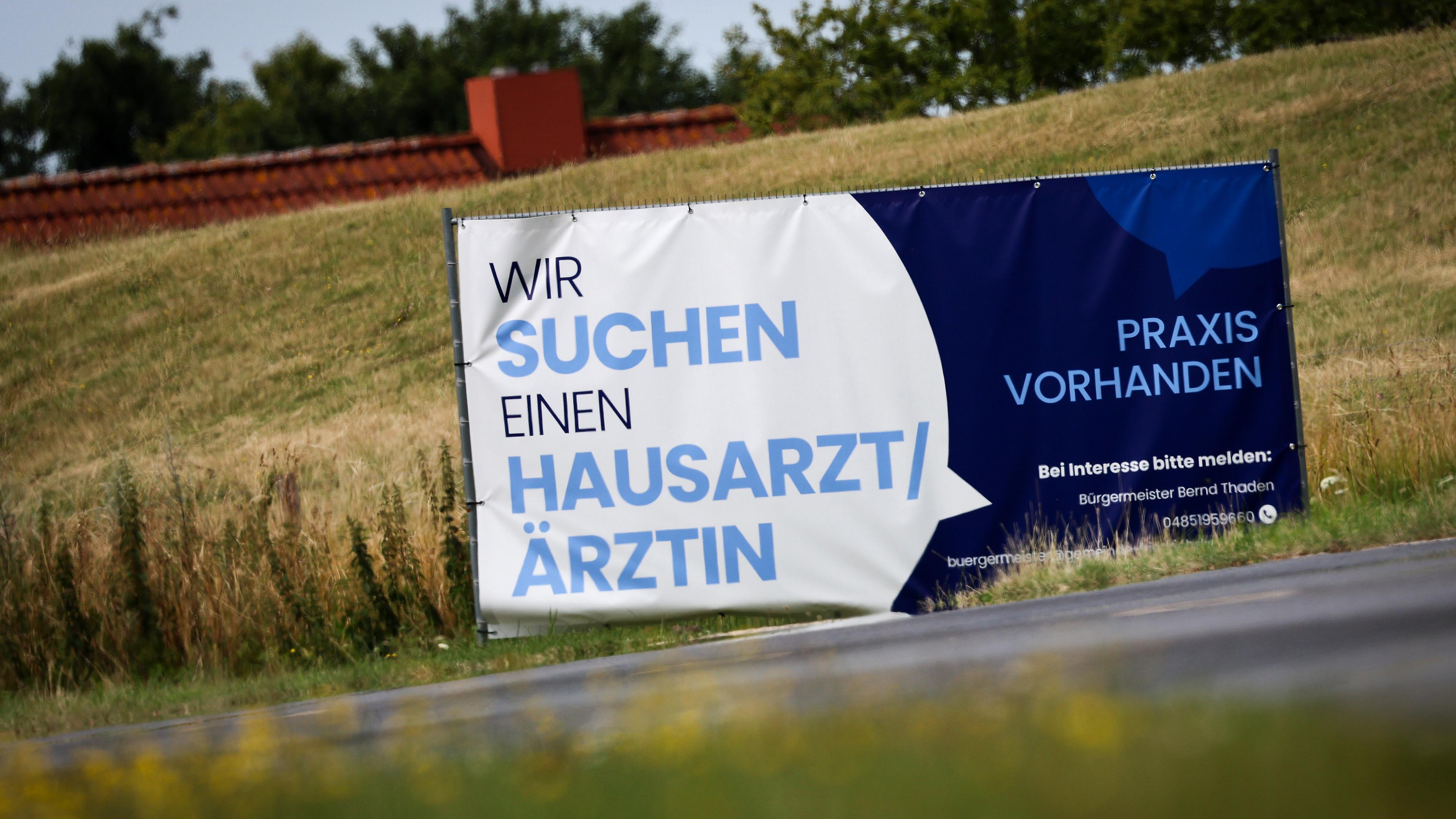 Ein Plakat mit dem Text "Wir suchen einen Hausarzt / Ärztin - Praxis Vorhanden" steht am Ortseingang von Friederichskoog