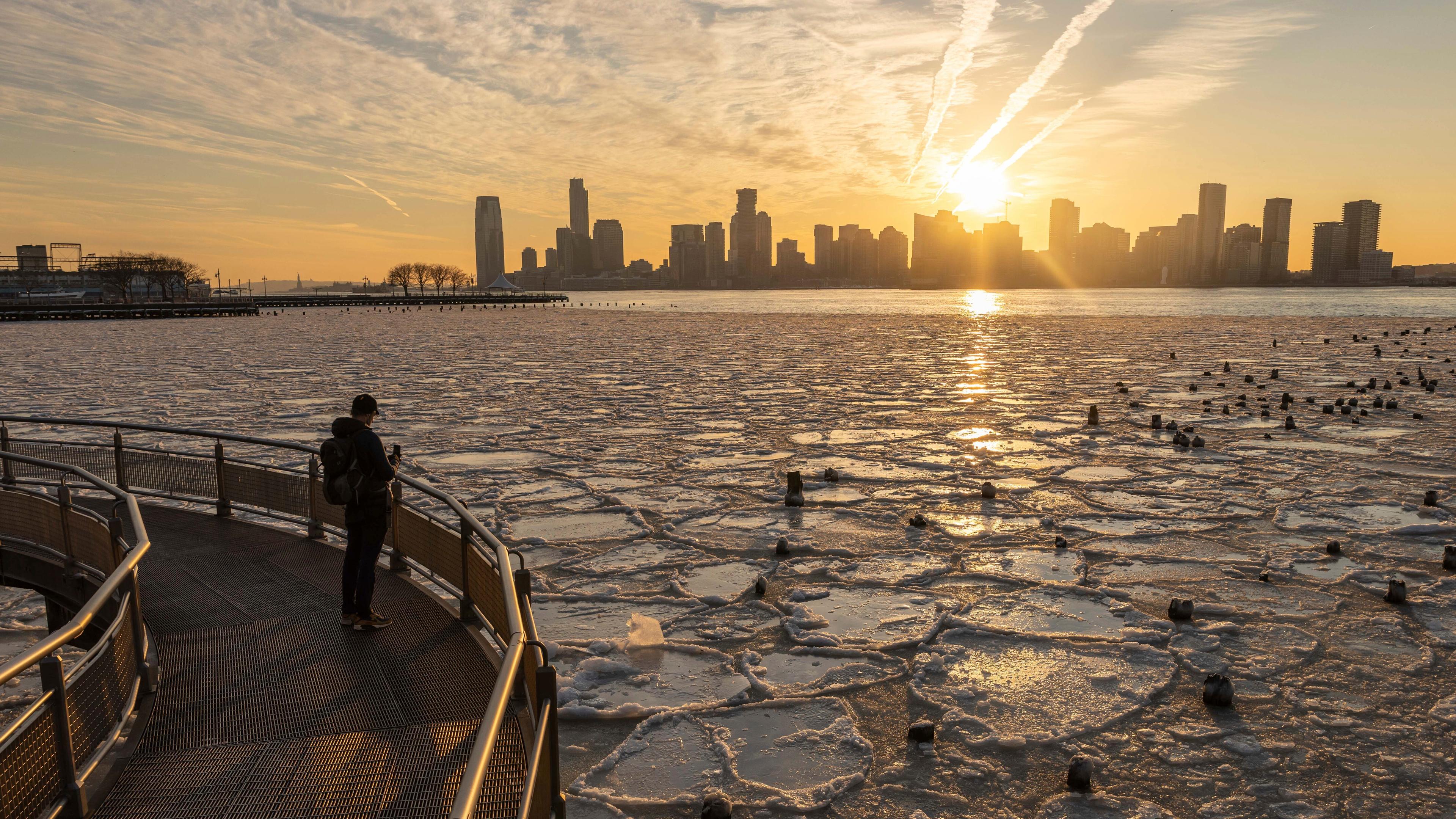 Der Hudson River bei Sonnenuntergang. Im Hintergrund sieht man die Skyline von New York. Ein Mann steht auf einer Brücke und fotografiert die mit vielen kleinen und großen Eisschollen bedeckte Flussoberfläche.