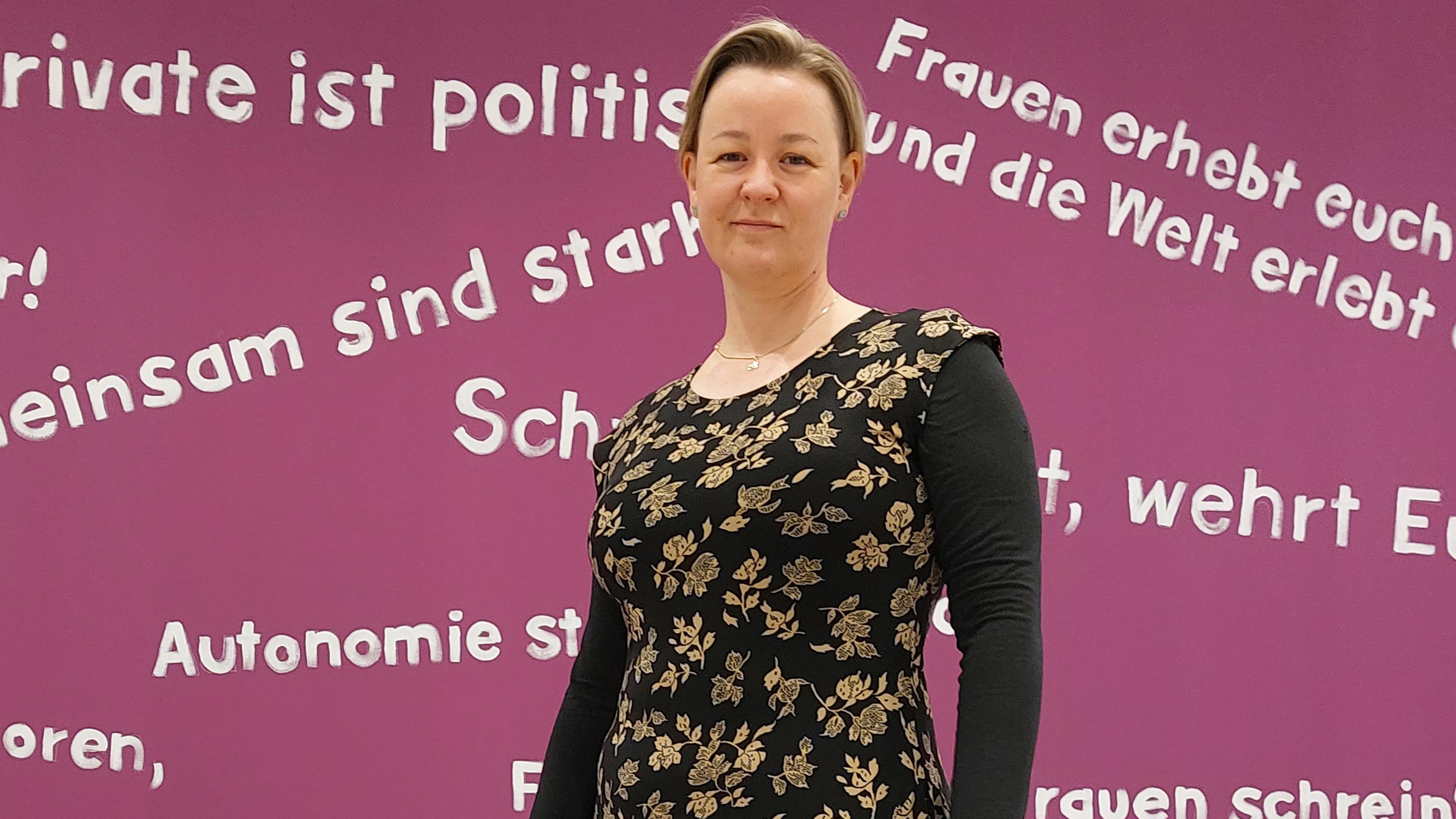 Sandra Schmitz vor beerenfarbener Wand mit diversen feministischen Schriftzügen.