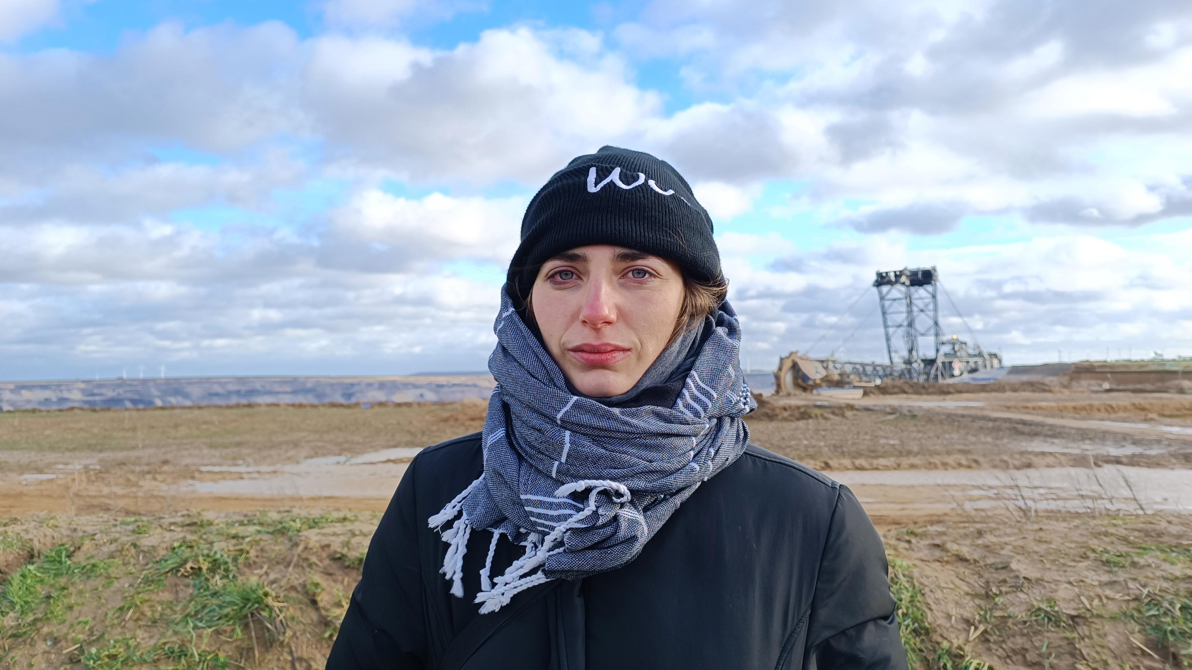 Junge Frau in Mütze und Schal gehüllt blickt ernst in die Kamera und steht vor dem weiten, leeren Gelände von Lützeratz. Im Hintergrund ein Braunkohlebagger zu erahnen, blauer Himmel mit zahlreichen Wolkenformationen.