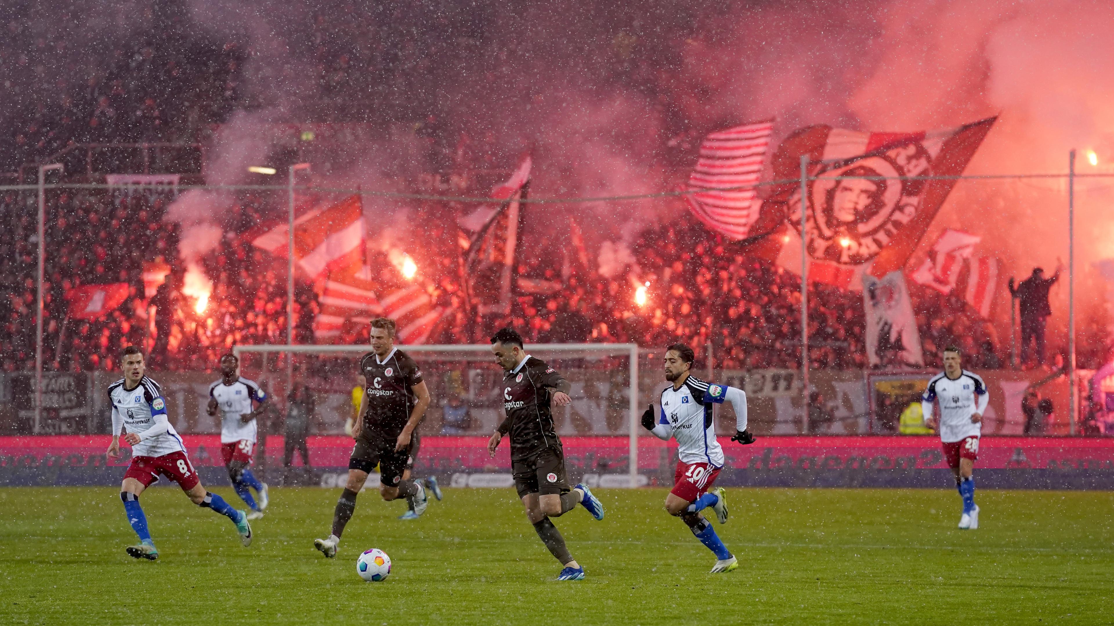 Im Vordergrund spielen mehrere Fußballspieler in weißen oder schwarzen Trikots auf einem Fußballplatz. Im Hintergrund stehen Fußballfans mit wehenden Fahnen und brennenden Böllern auf der Tribüne, die dadurch rötlich beleuchtet wird.