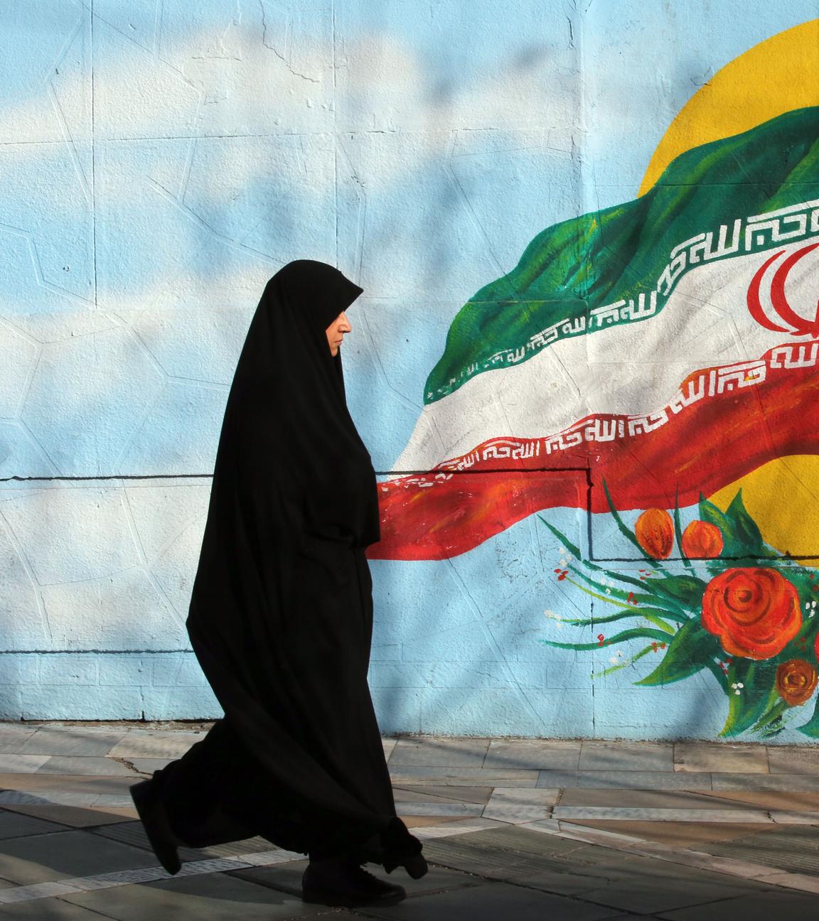 Frau mit Kopftuch läuft vor einer Wand, wo eine iranische Flagge zu sehen ist