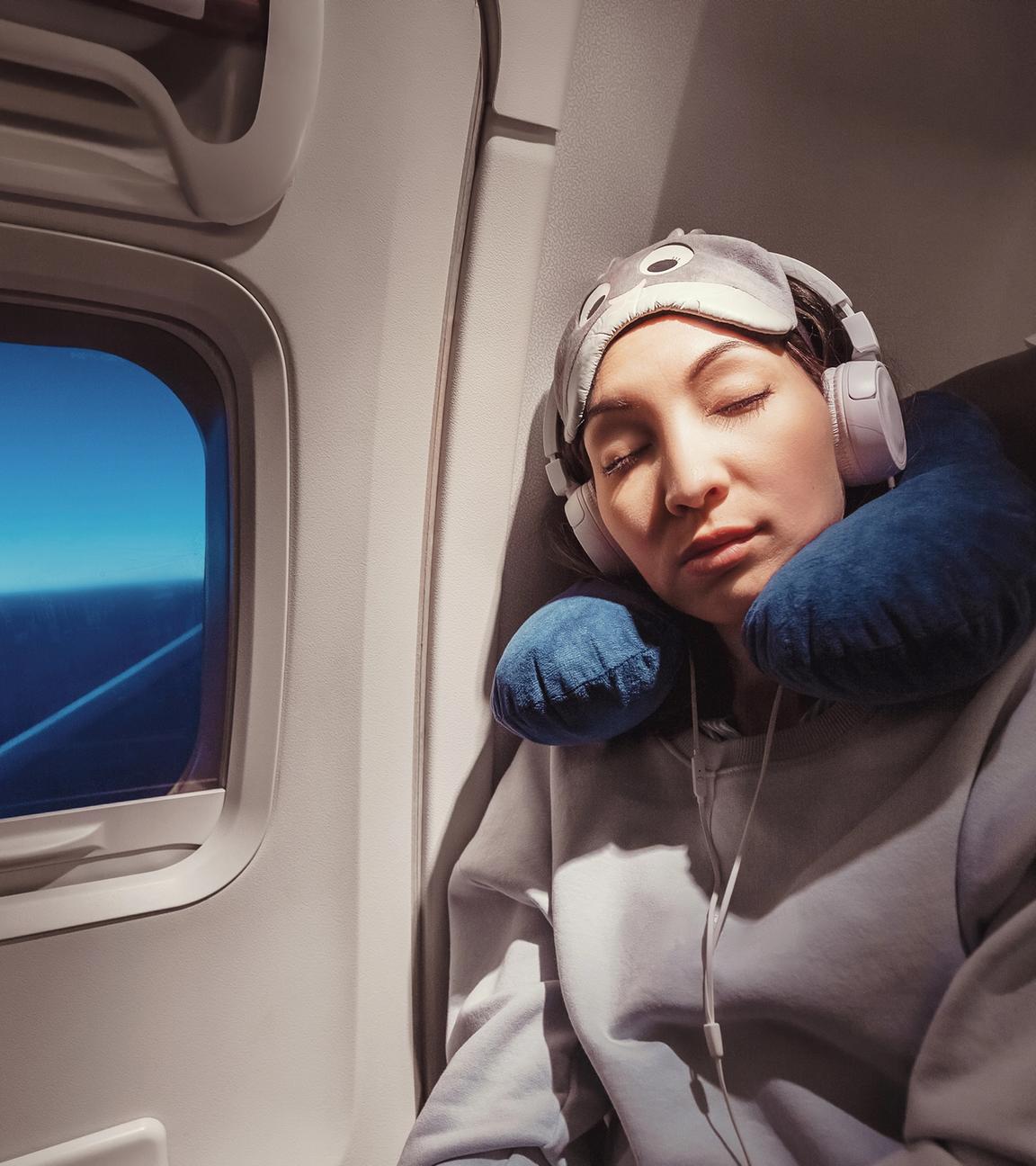 Frau mit Kopfhörern und Nackenkissen sitzt im Flugzeug und schläft.