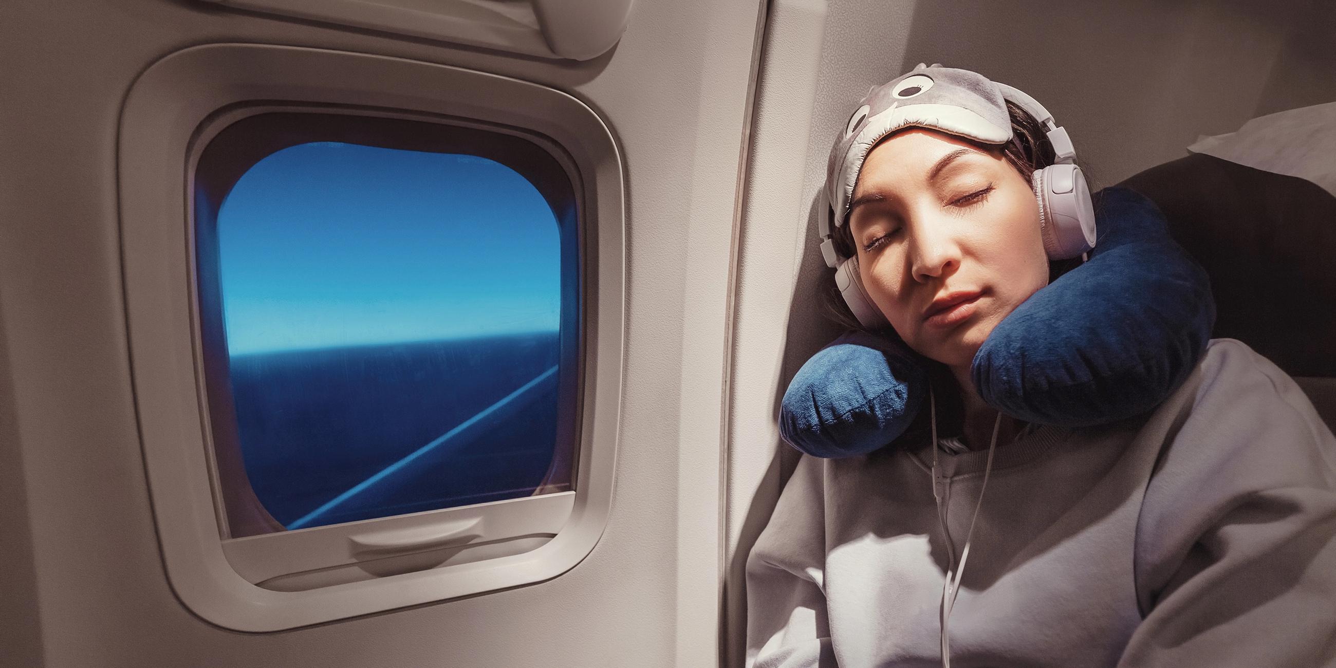 Frau mit Kopfhörern und Nackenkissen sitzt im Flugzeug und schläft.