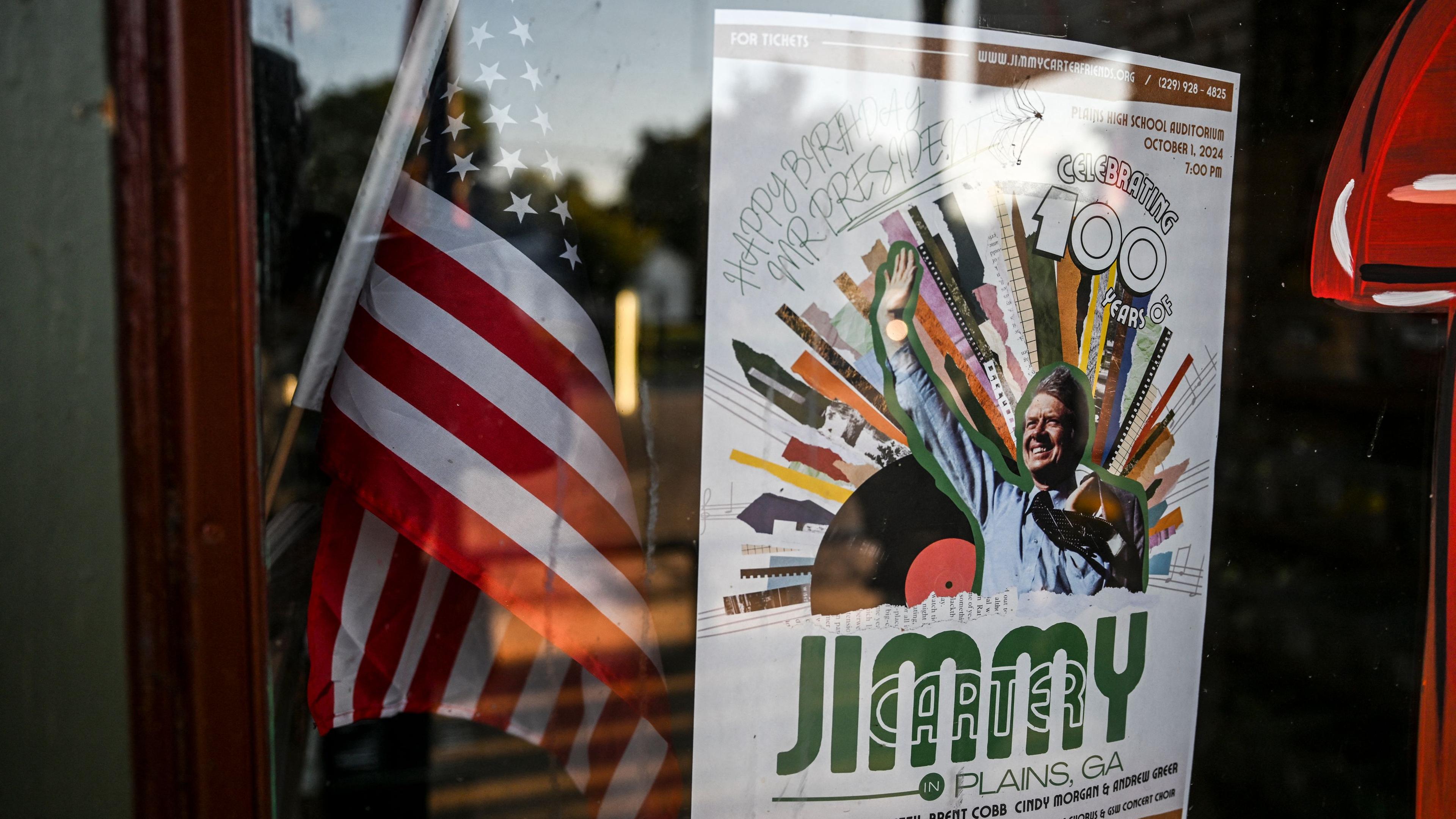 Die US-Amerikanische Flagge hinter einer Schaufensterscheibe. Daneben ein Plakat zu Ehren des hundertsten Geburtstag vom ehemaligen Präsidenten der Vereinigten Staaten: Jimmy Carter.