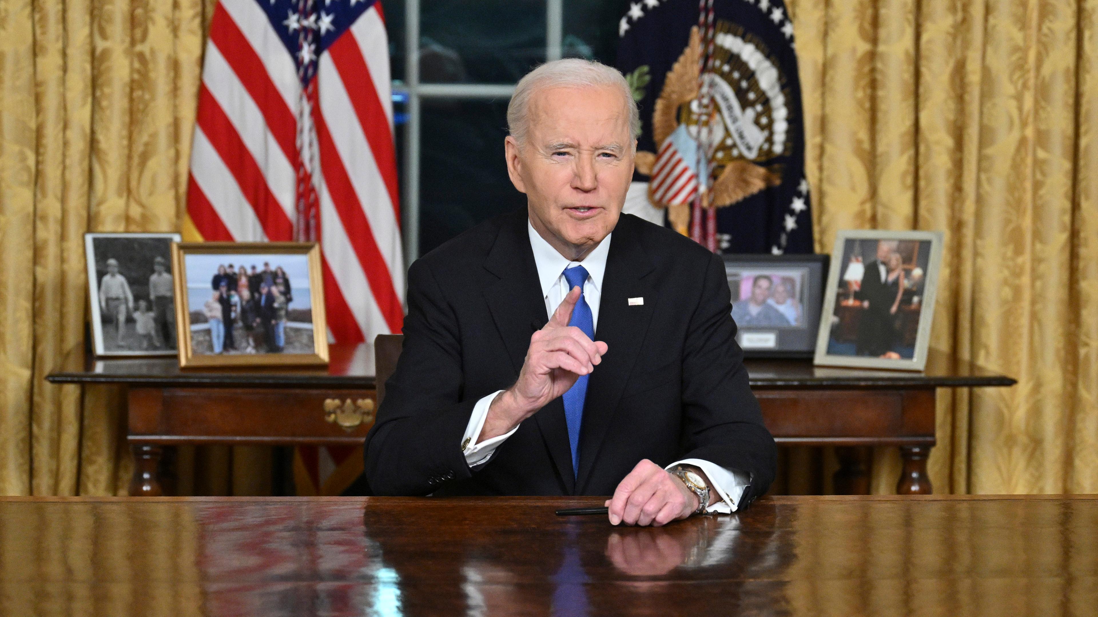 Joe Biden sitzt im Oval Office hinter dem Resolute Desk und spricht mit erhobenem Zeigefinger in die Kamera. Hinter ihm stehen Familienfotos.