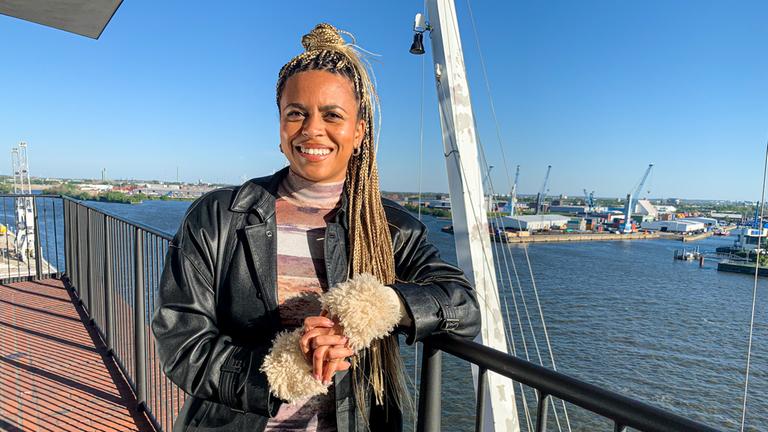 Aminata steht auf dem Balkon der Elbphilamonie. Im Hintergrund liegt der Hafen mit mehreren Kränen.