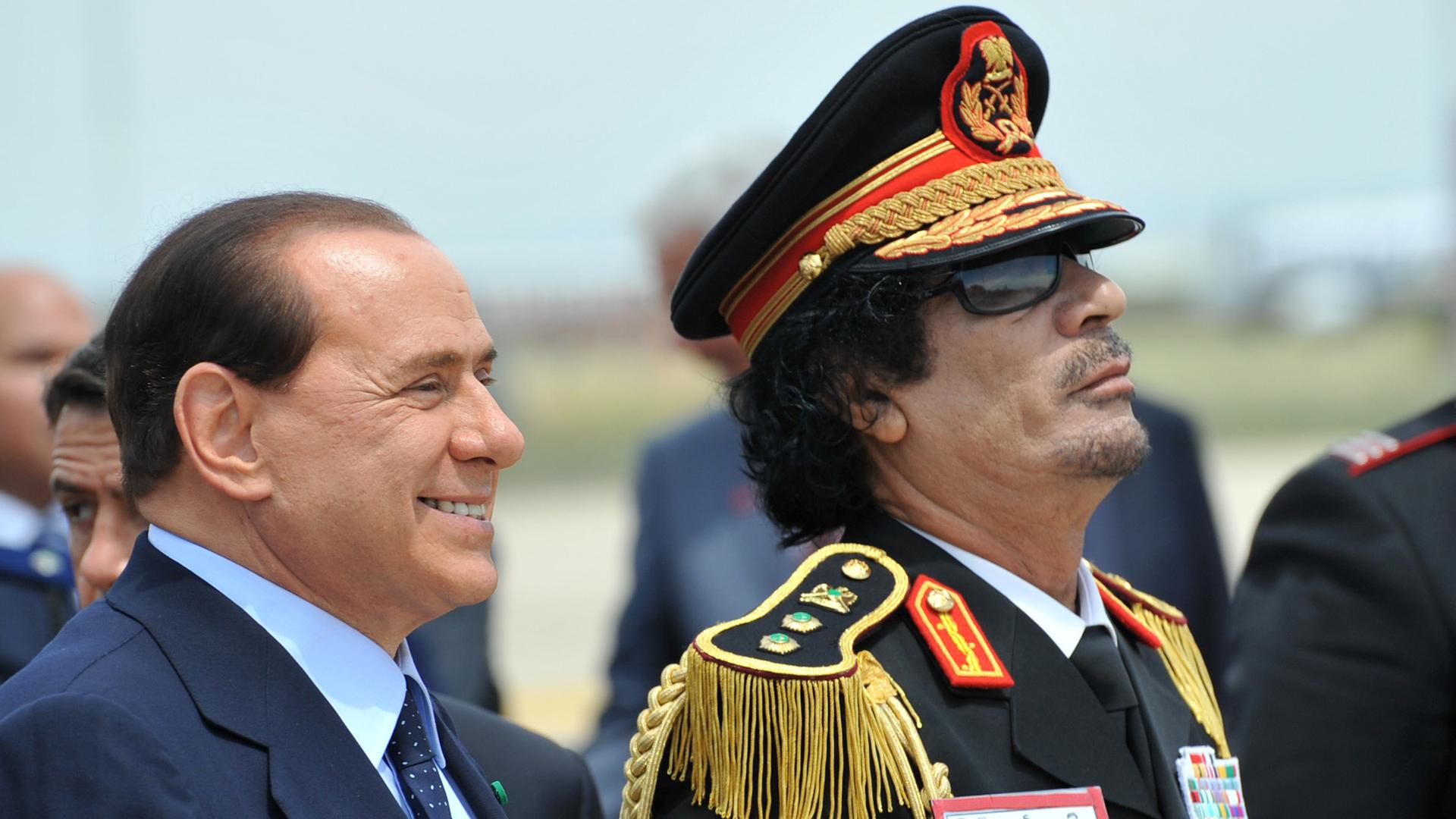 "Killing Gaddafi - Jagd auf den Diktator": Nahaufnahme von Gaddafi und Berlusconi. Sie stehen nebeneinander und schauen in eine Richtung.