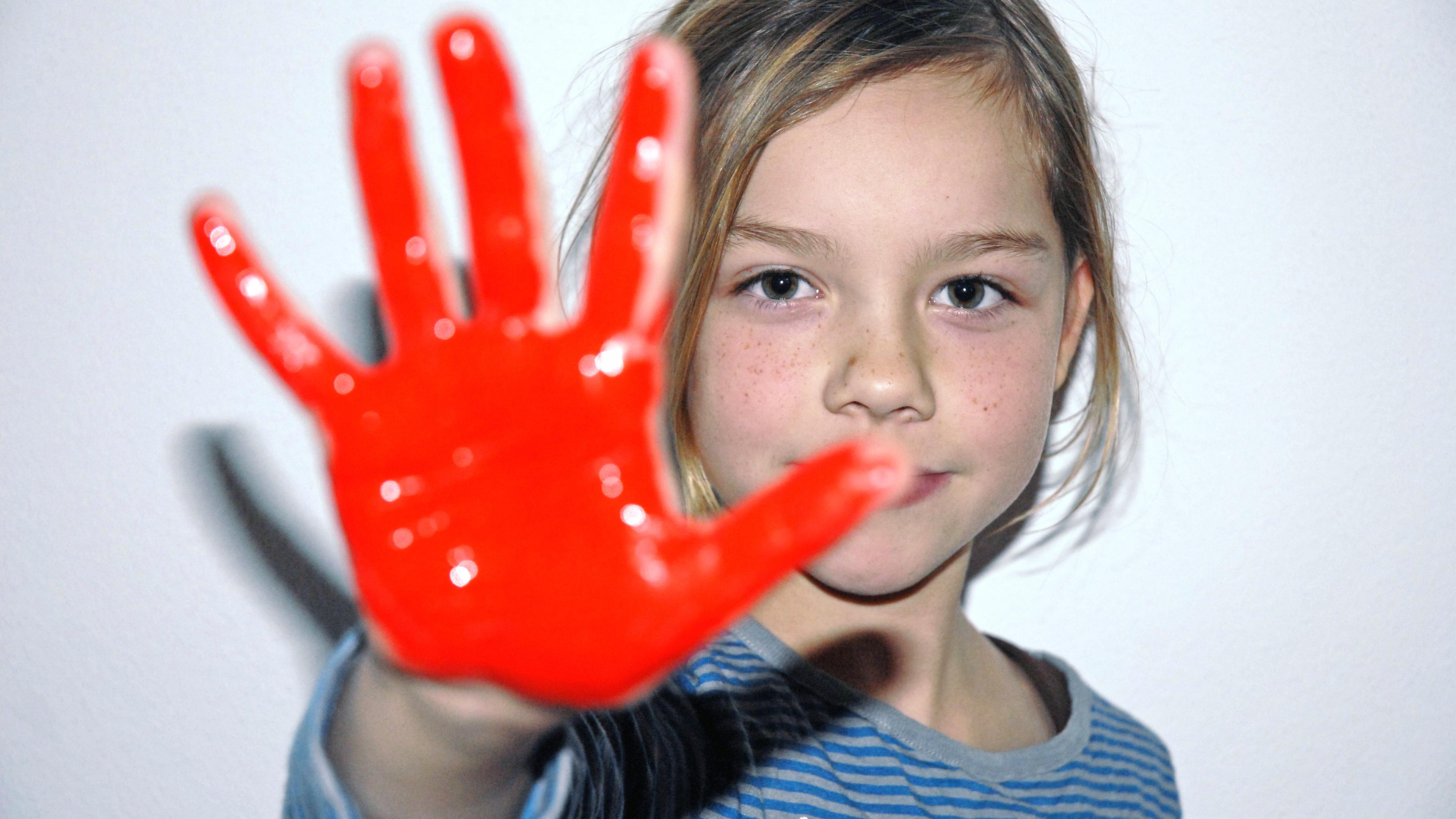 Ein Kind streckt eine Hand, die rot angemalt ist, in die Kamera. Sie hat blonde Haare. 